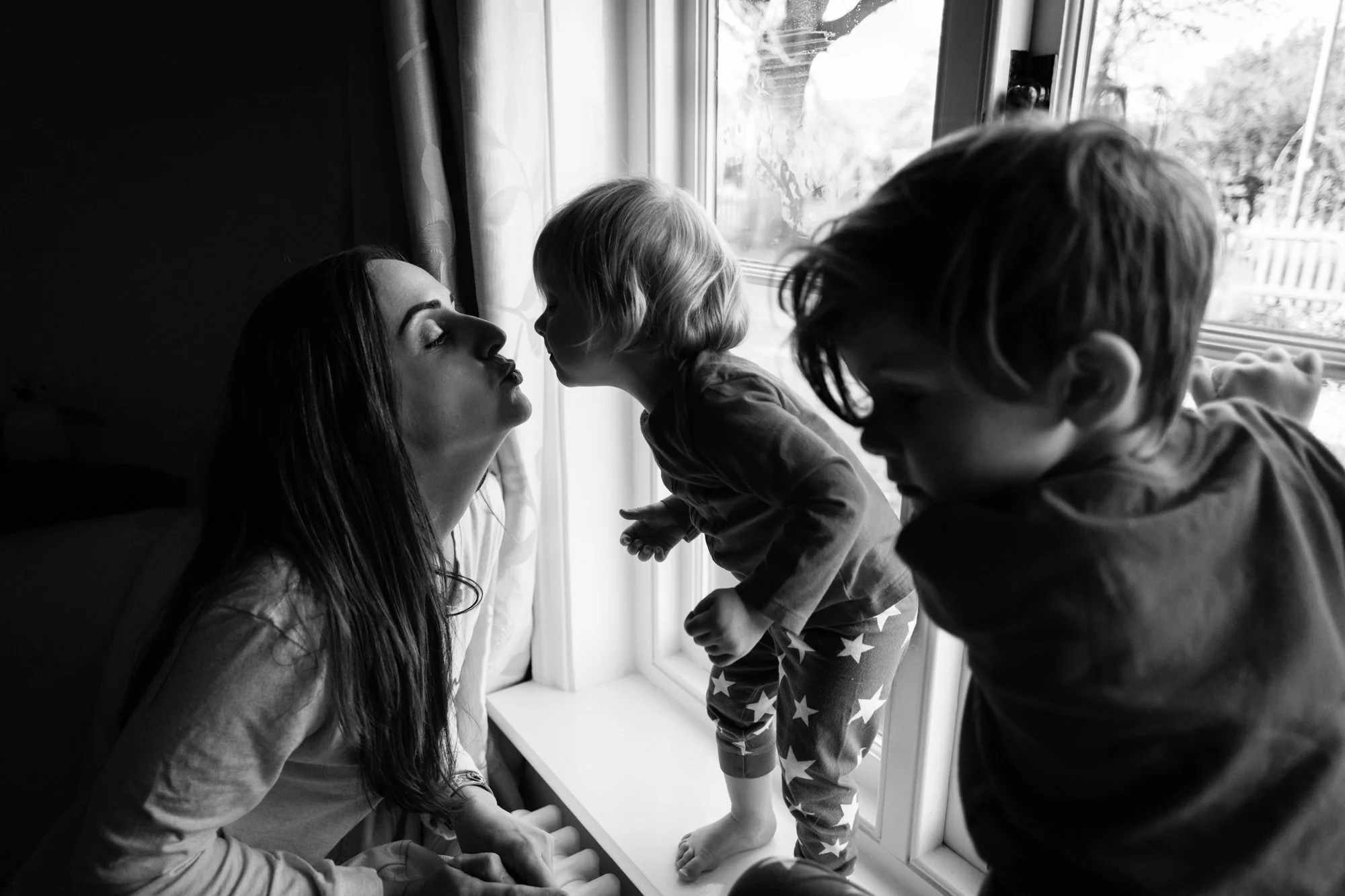 A woman with long dark hair and two young children, a girl and a boy, near a window with curtains. The girl is standing on the windowsill, leaning forward as if to kiss the woman, while the boy is sitting close to her, both children leaning toward he