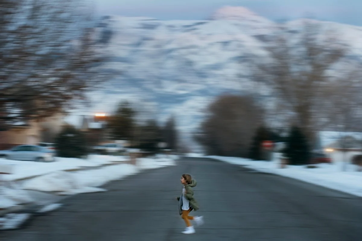 A young girl running on a snow-covered suburban street with mountains in the background during winter.