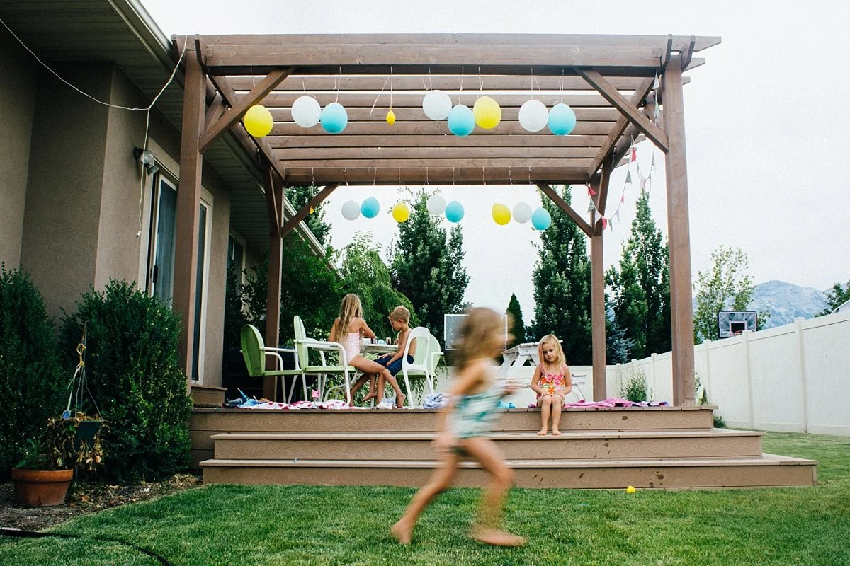Children playing on a backyard deck decorated with hanging paper lanterns, with a table and chairs, and a white fence surrounding the yard.