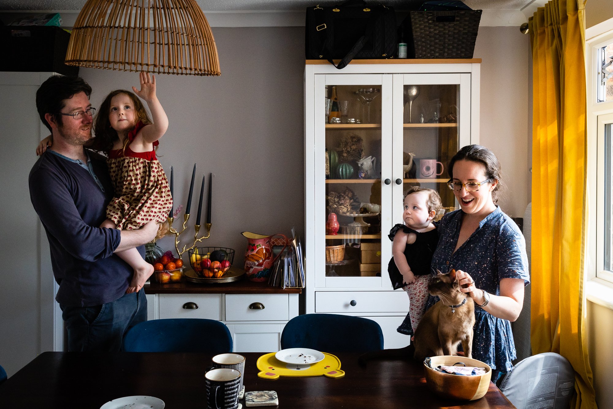 A family enjoying a moment in their dining room. A man is holding a young girl, and a woman is holding a smaller child and pet dog. The room has yellow curtains and a white cabinet with glass doors.