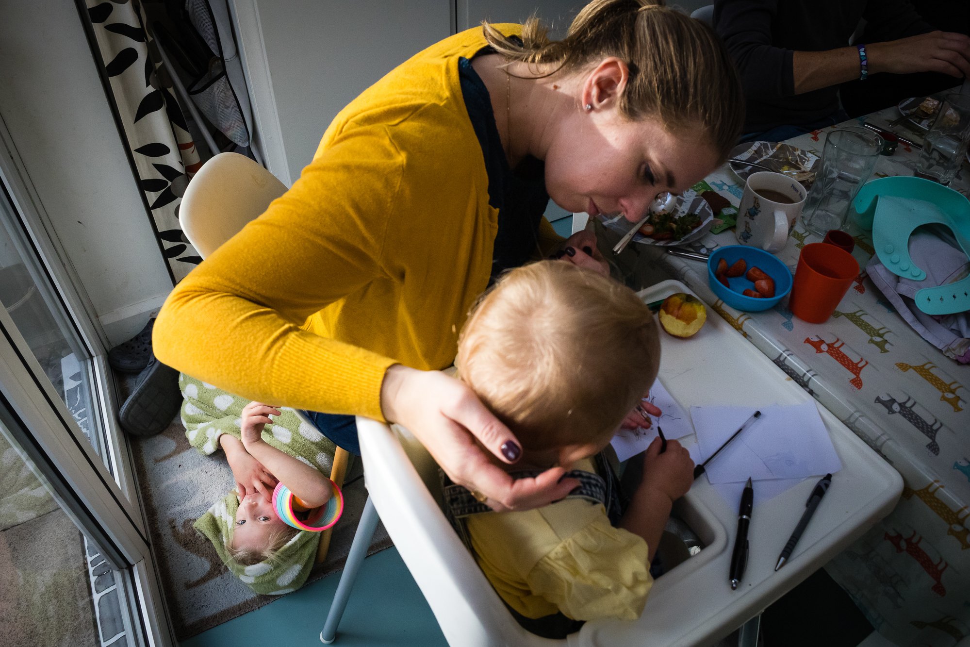 A woman leaning over a young child sitting in a high chair, with another small child lying on the floor nearby. The table has snacks, cups, and pens.