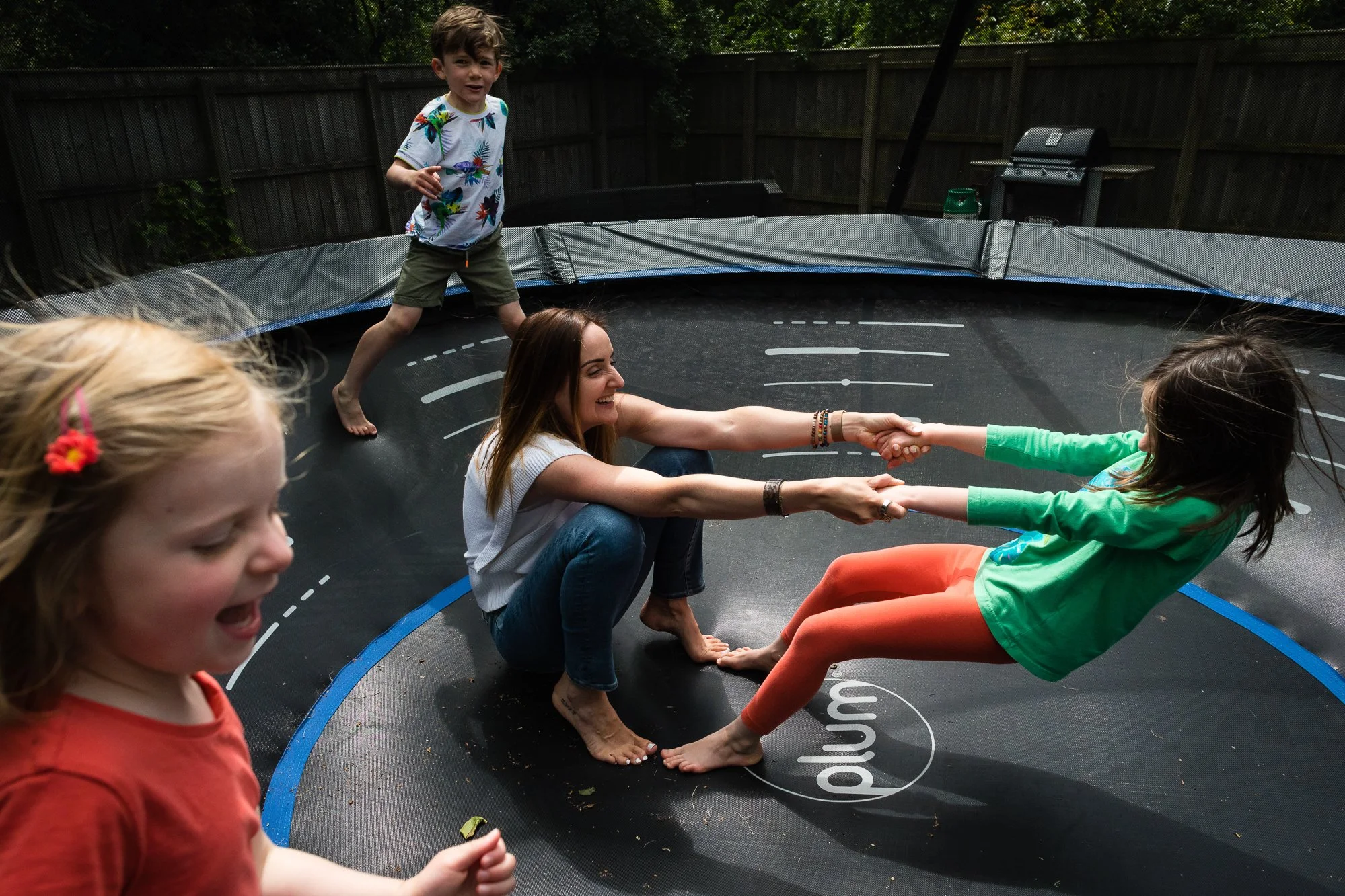 A woman and a girl are sitting on a trampoline holding hands while the girl leans back. Two children are nearby, one standing and one smiling at the camera.