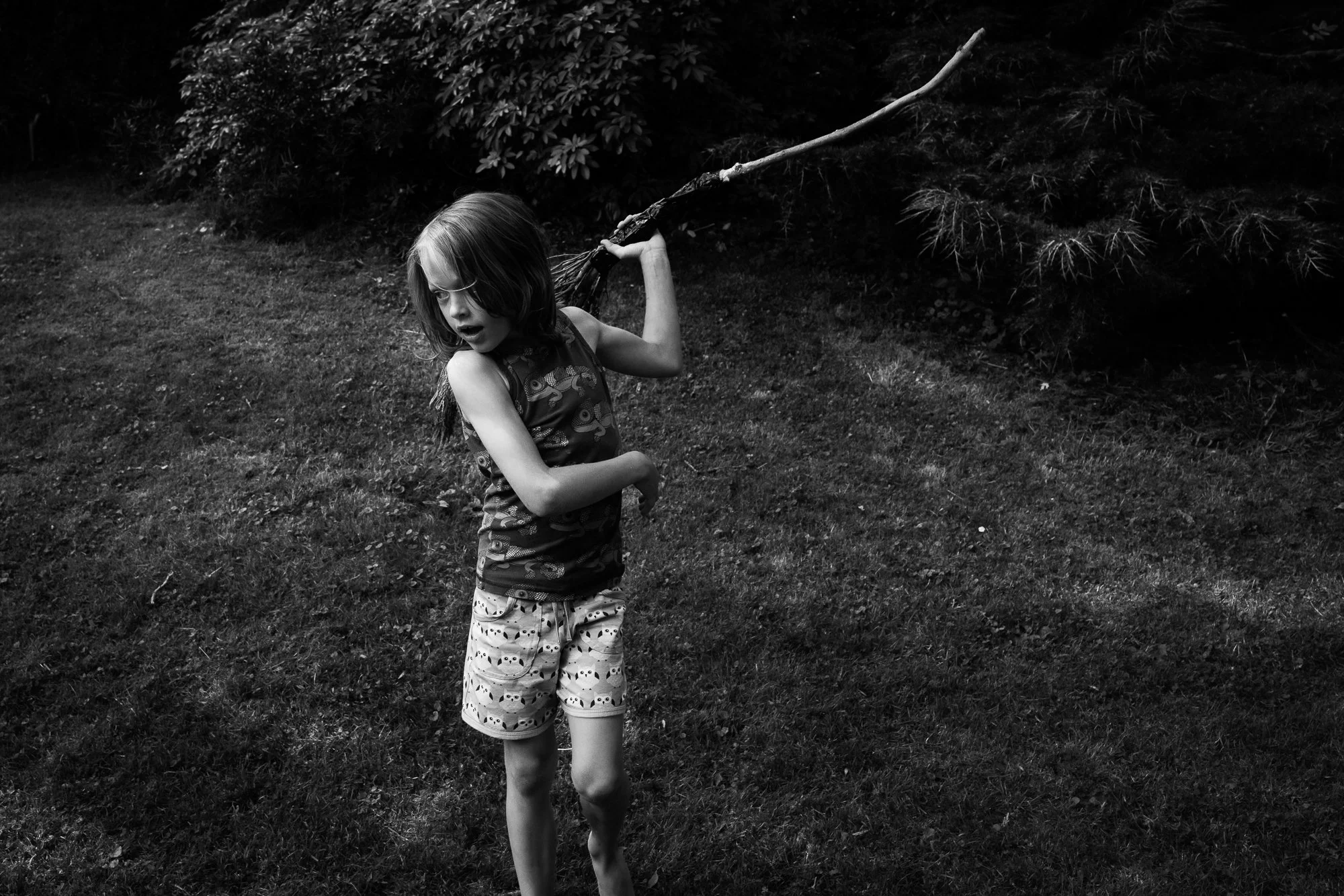 A young girl with shoulder-length hair, wearing a patterned sleeveless top and shorts, holding a large stick over her shoulder in an outdoor grassy area with trees.