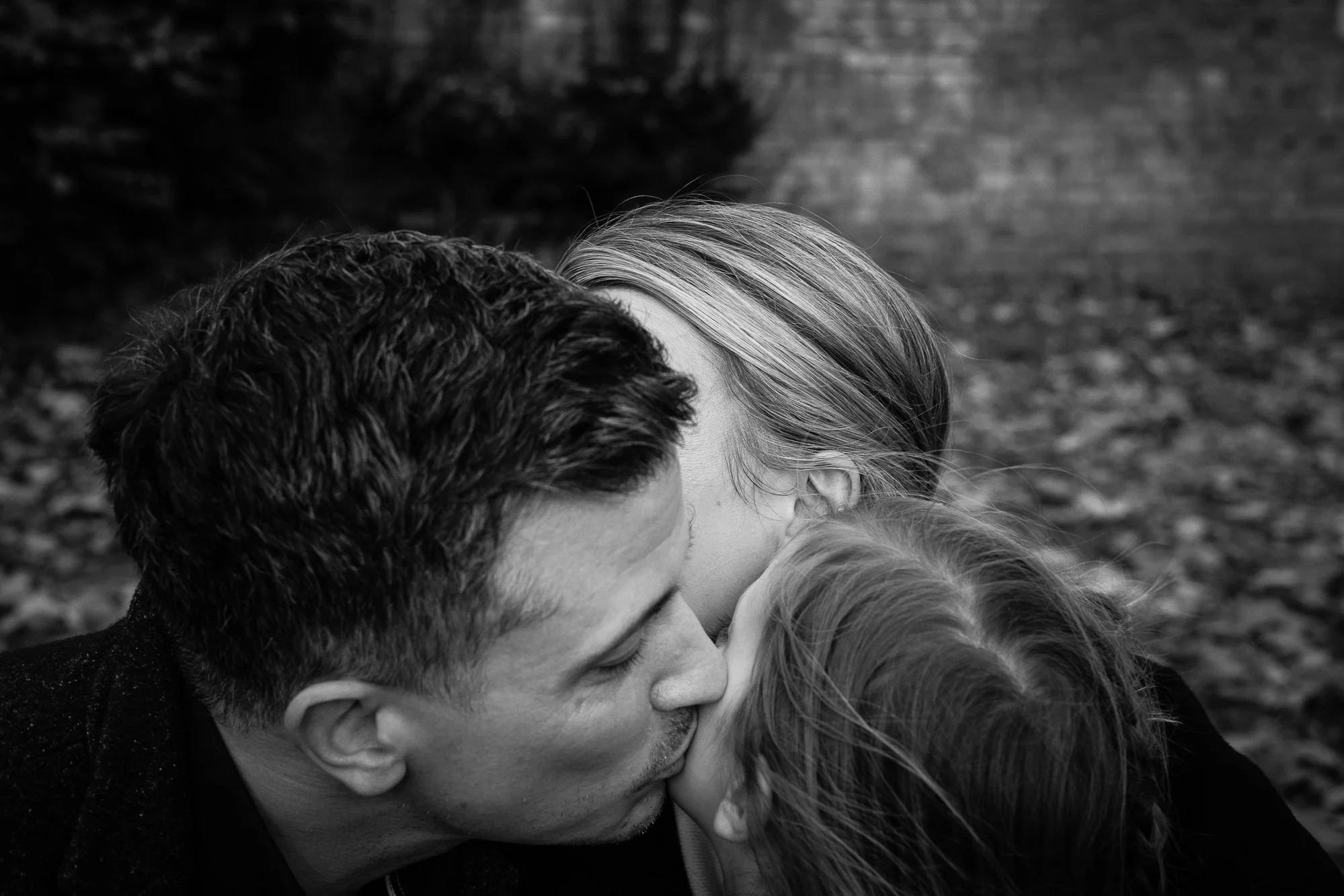 A black and white photo of a couple kissing, with a child in front of them. The scene takes place outdoors, with fallen leaves on the ground.