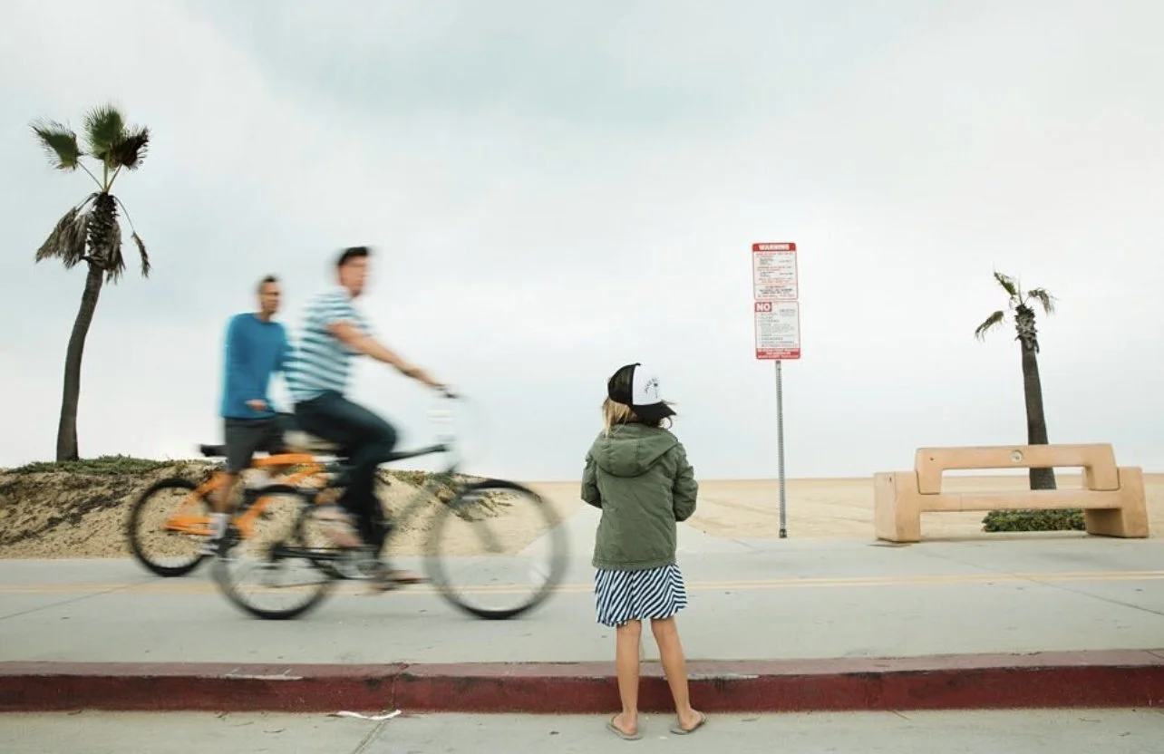 A girl with a helmet and striped dress standing on sidewalk watching two blurred bikers riding past near the beach, with palm trees, a red and white sign, and a bench in the background.