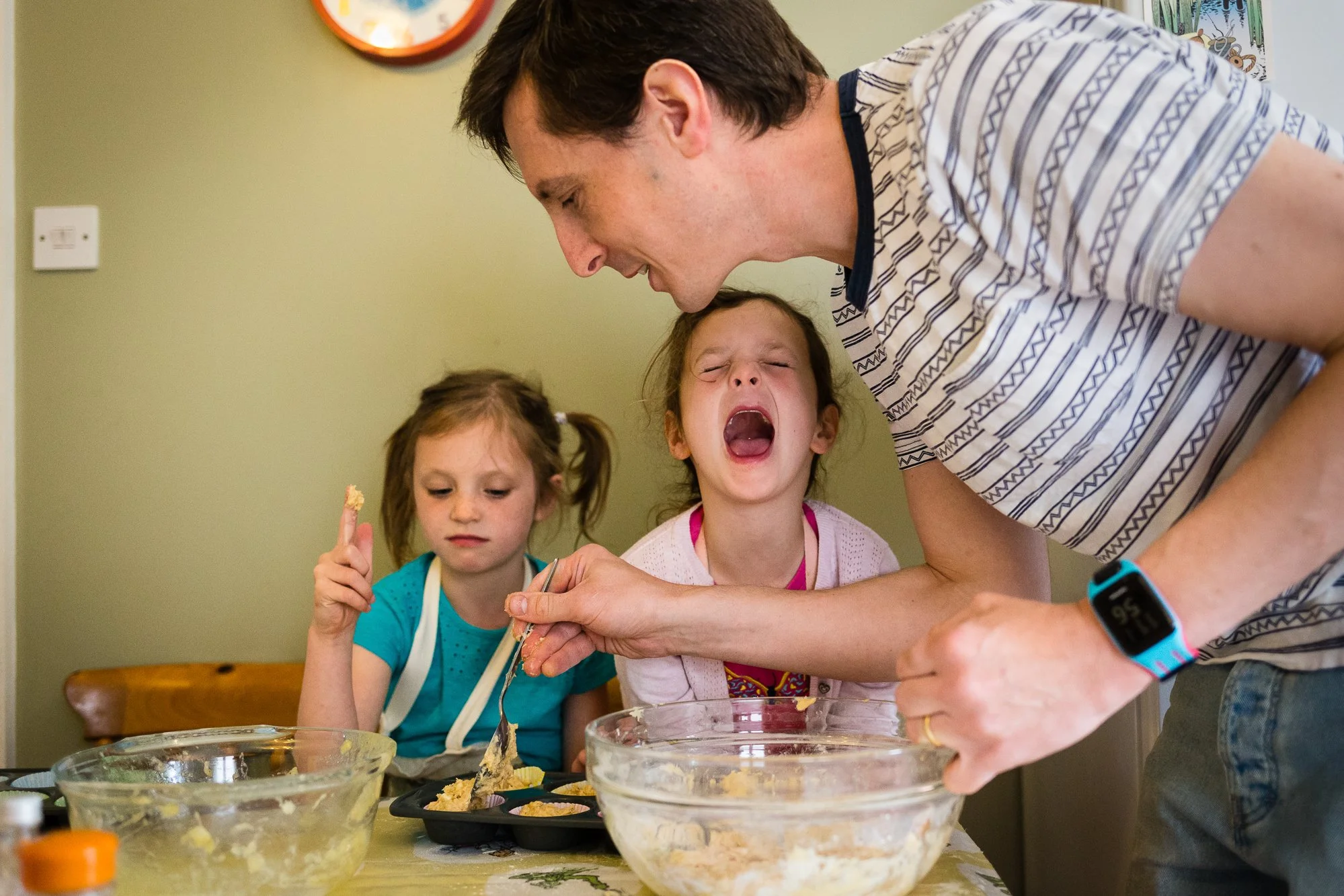 Man teaching children to bake or cook in a kitchen, with one girl yawning and another girl holding a spoon with batter.