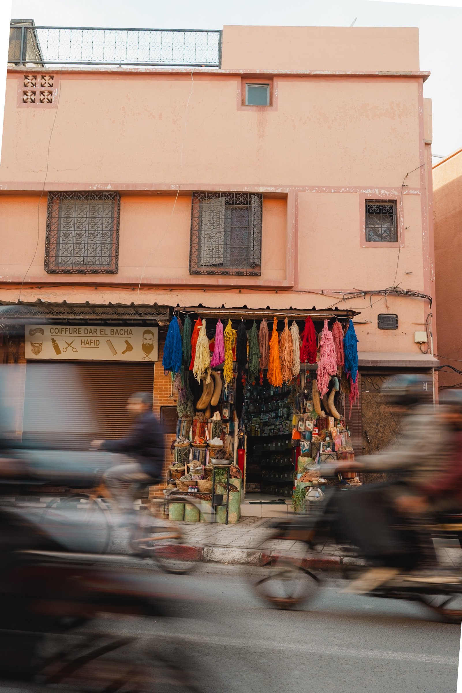 Street view of a pink building with a storefront selling textiles and accessories. Colorful skeins of yarn hanging above the entrance, with shoes and small items displayed on racks at the entrance. Two motorbikes with blurred motion pass in front, in