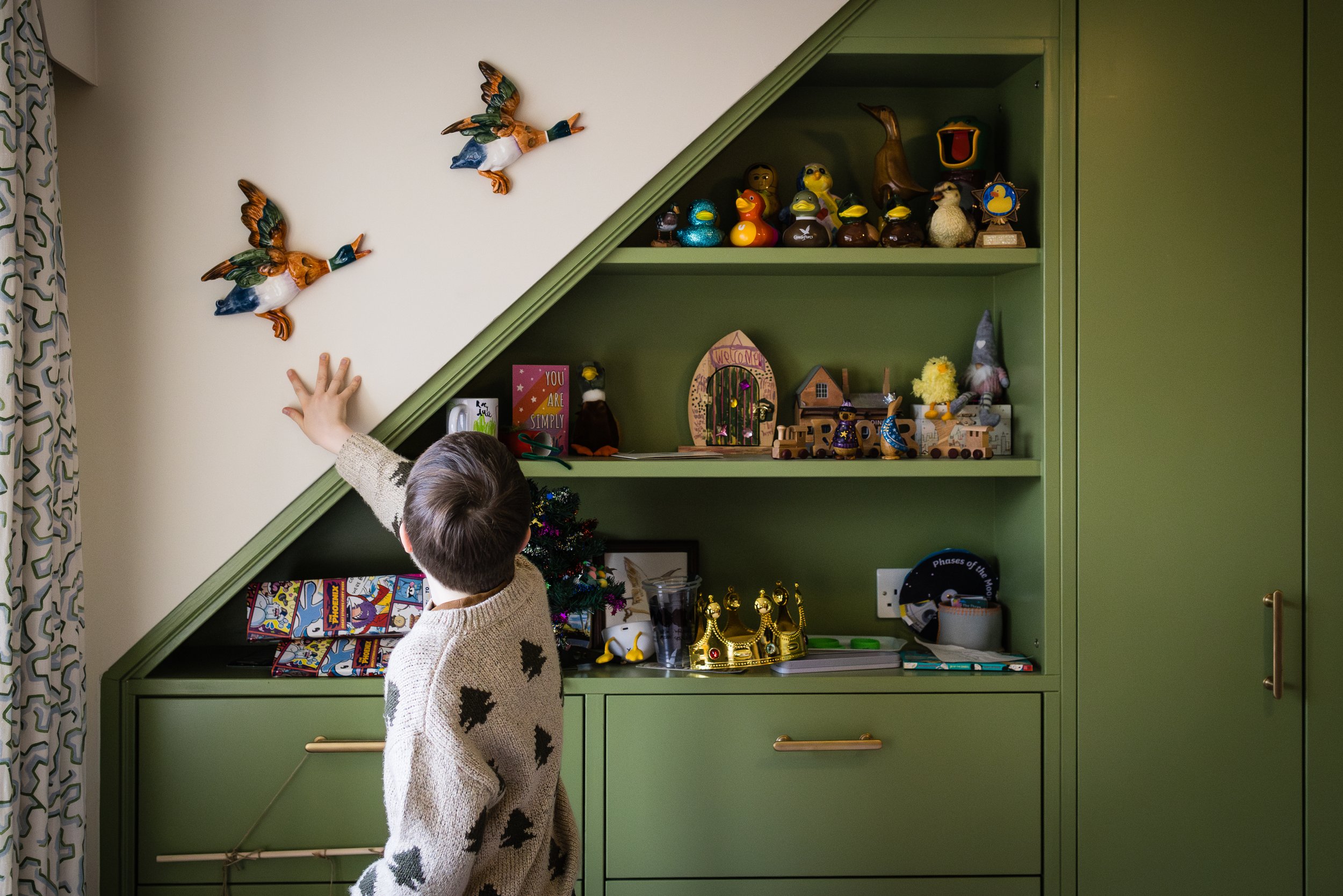Child reaching up towards decorative ducks on a slanted green and white wall shelf in a cozy, decorated room.