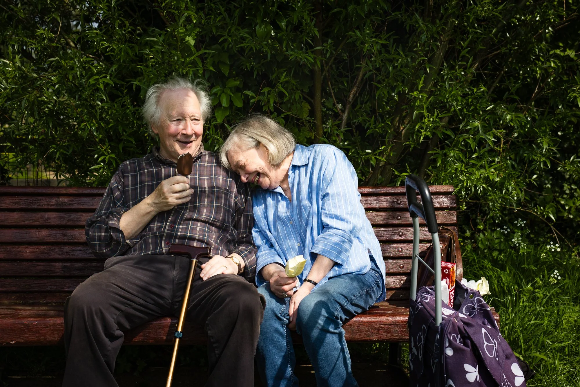 An elderly man and woman sit on a park bench, sharing a laugh while enjoying ice cream. The man holds a chocolate popsicle, and the woman has a cup of ice cream. They are surrounded by green foliage, and there is a cane resting against the bench and 