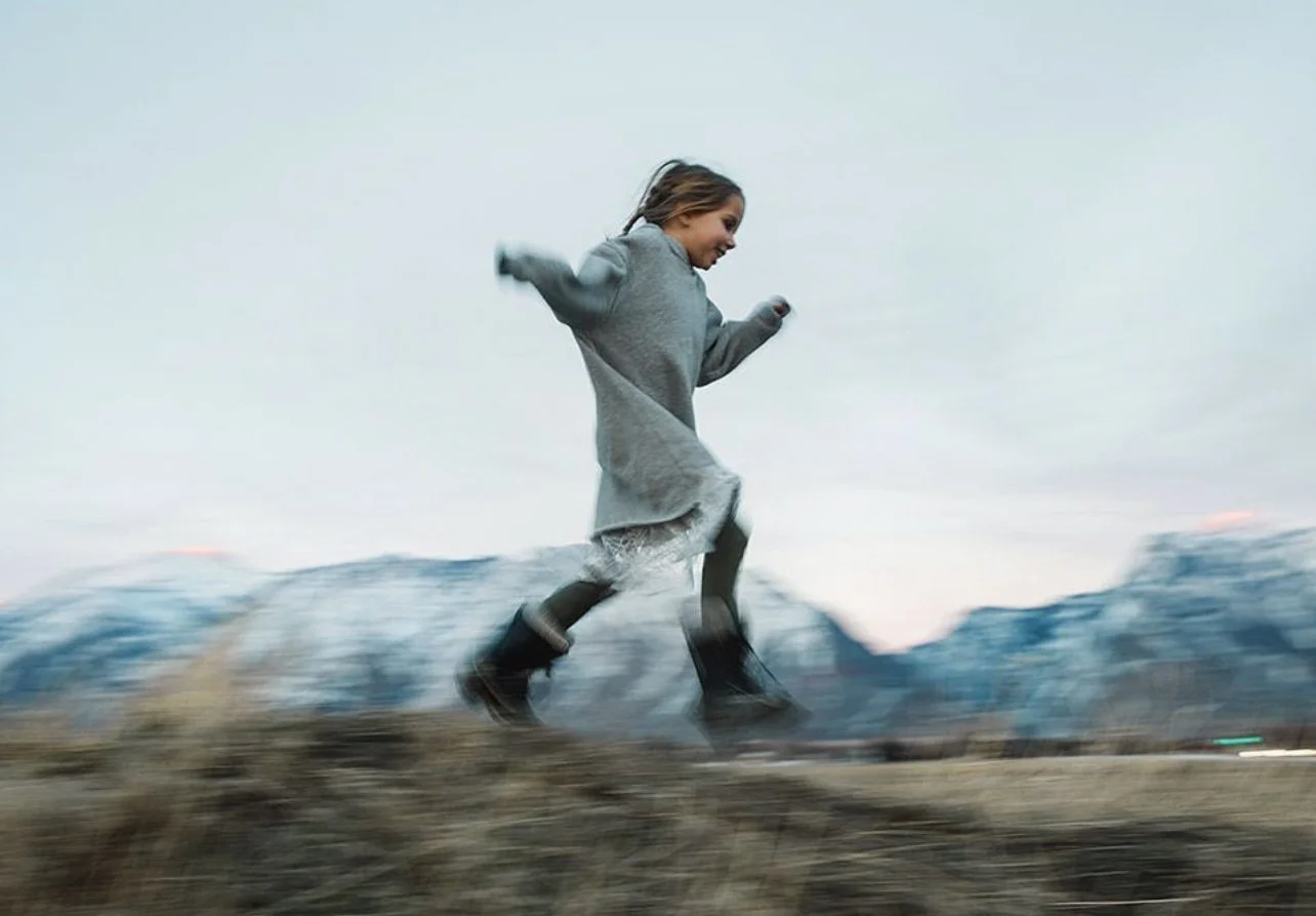 A young girl running outdoors in a mountainous area during cloudy weather, wearing a gray coat and black boots.