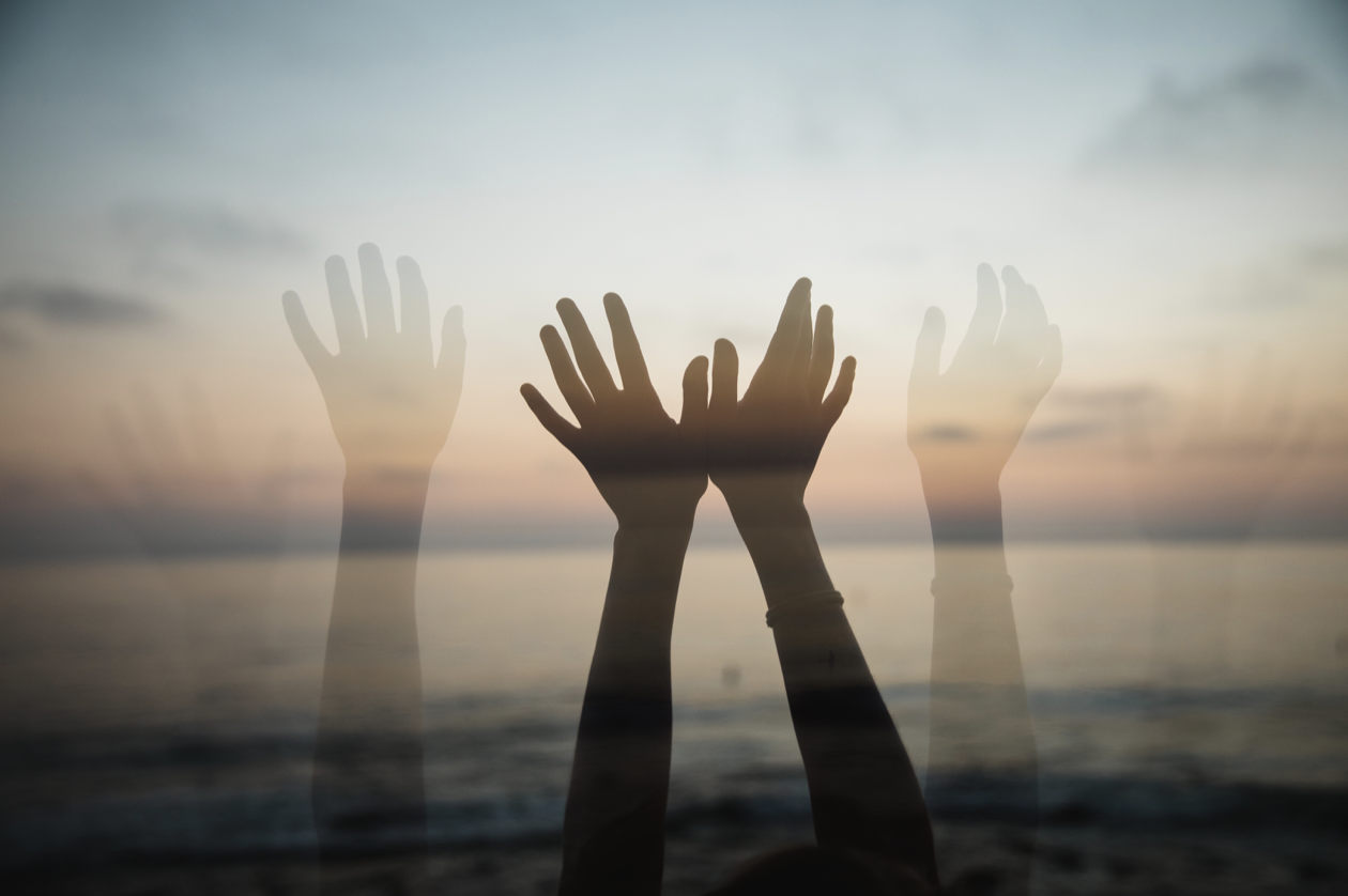 Multiple hands raised in front of a sunset over the ocean, creating a layered, transparent effect.