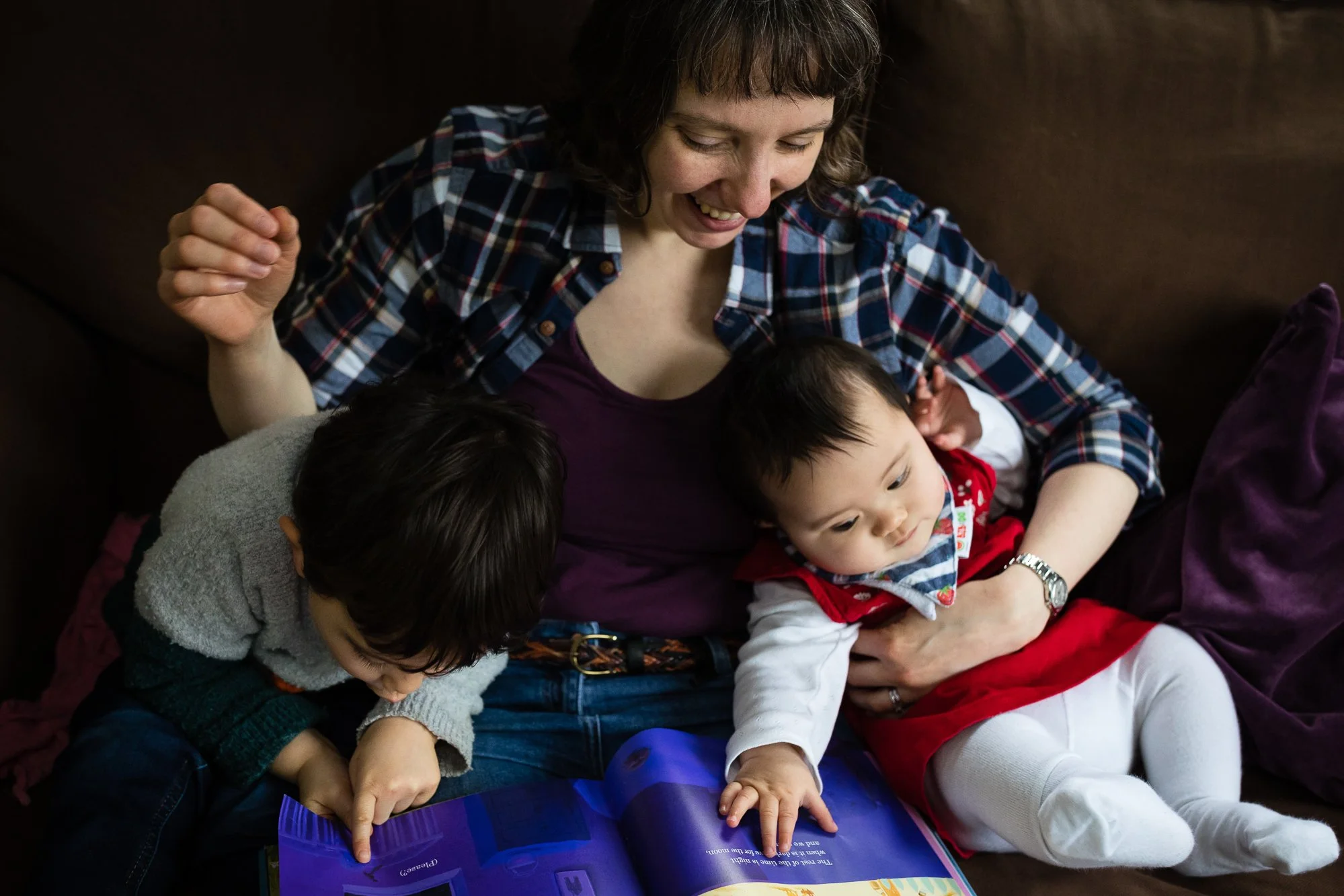 A woman sits on a couch with two young children, reading a colorful book together while smiling and engaging with them.