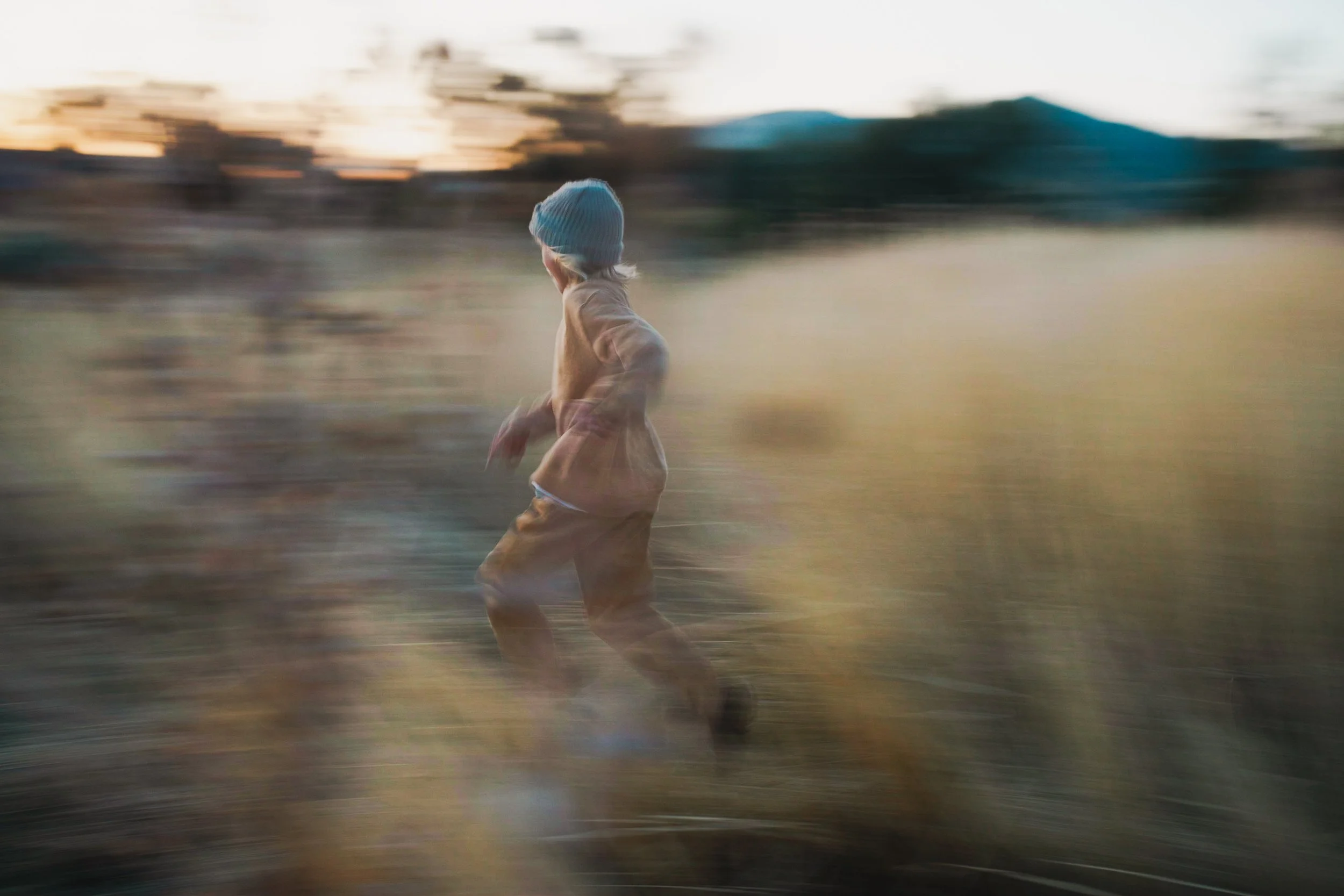 A young person running outdoors through a field with a blurry background during sunset, wearing a grey beanie, a beige jacket, and brown pants.