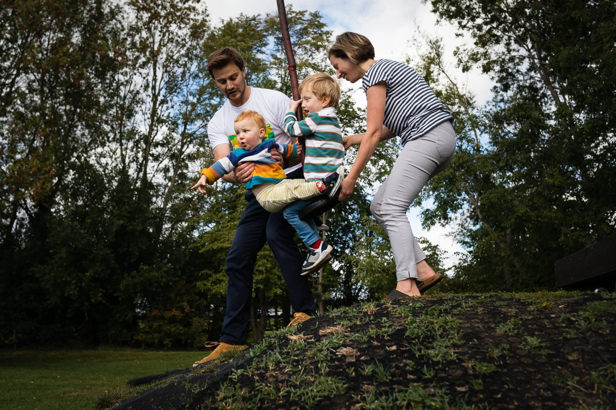 Family of four, including two small children, helping each other on a log in a park surrounded by trees.