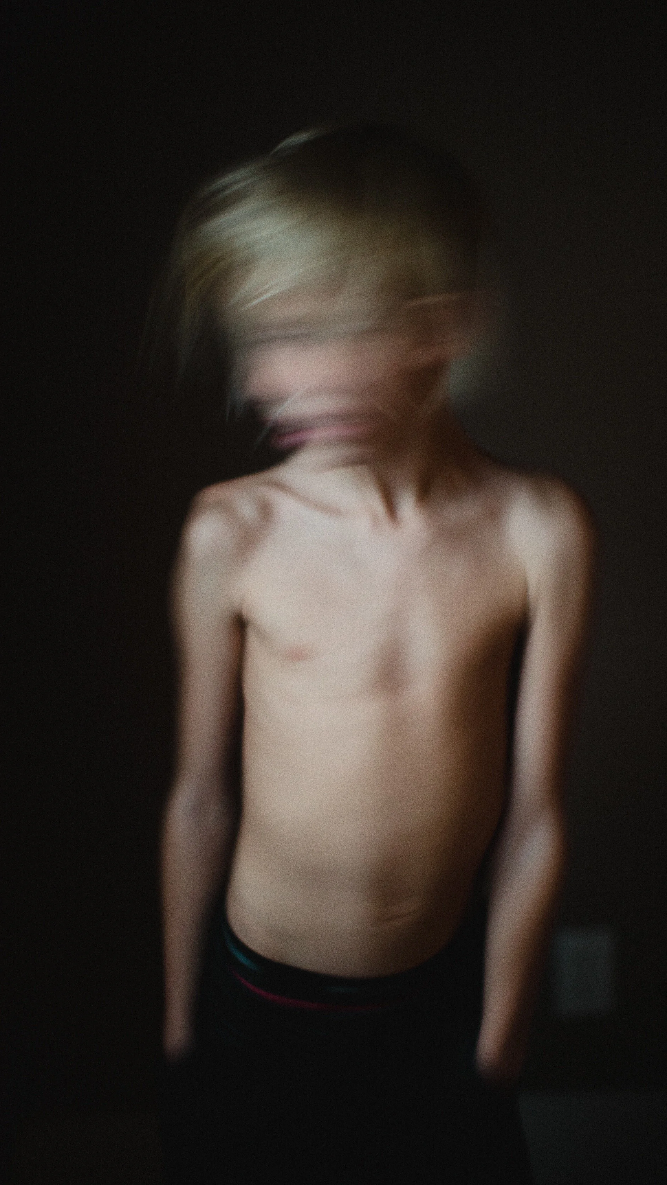 A blurred image of a shirtless person with short hair, standing against a dark background.