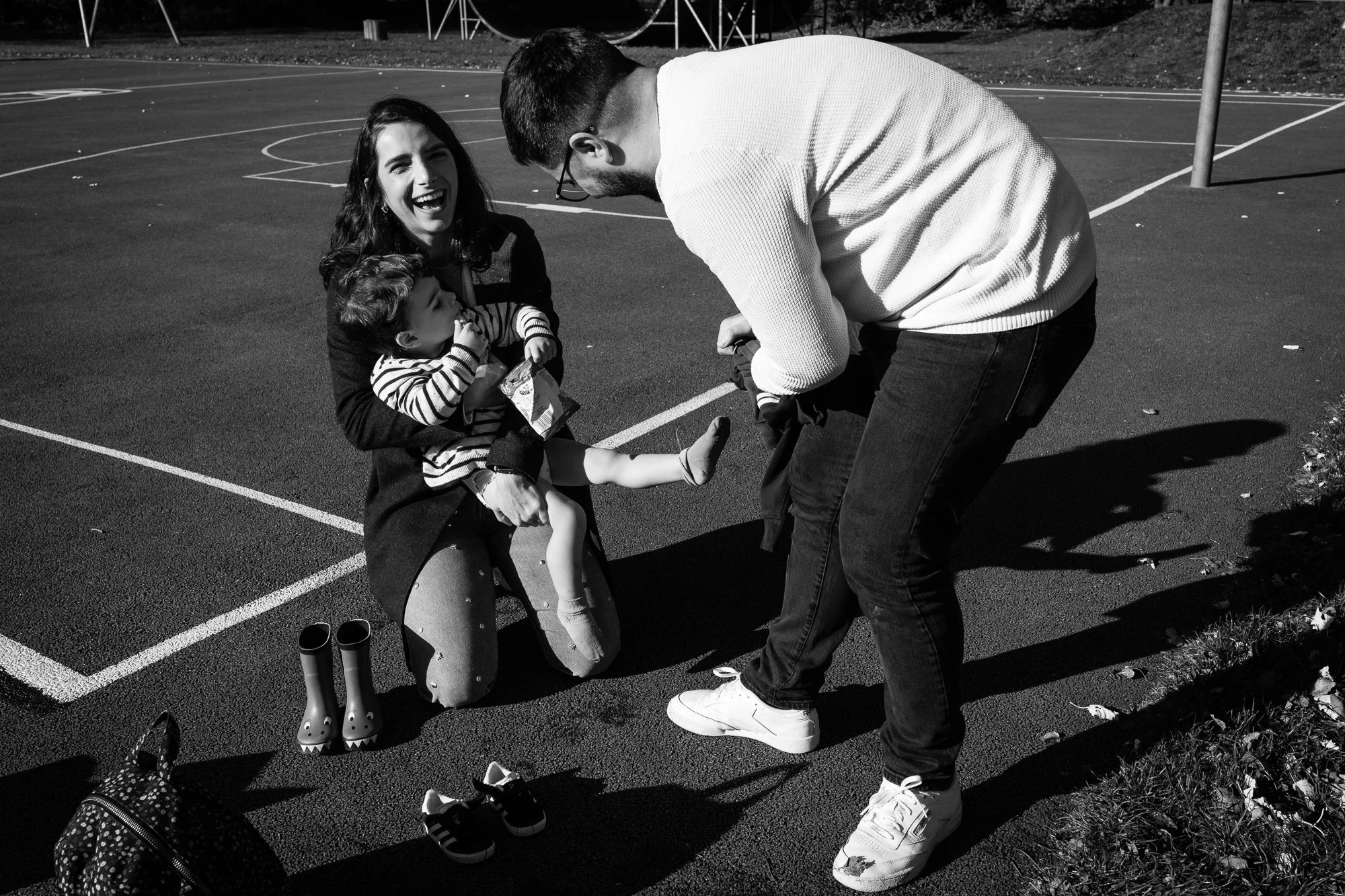 A woman kneeling on a basketball court, holding a young girl in her arms, laughing. A man in white sneakers bent over, smiling at the girl. There are shoes and a backpack on the ground nearby.