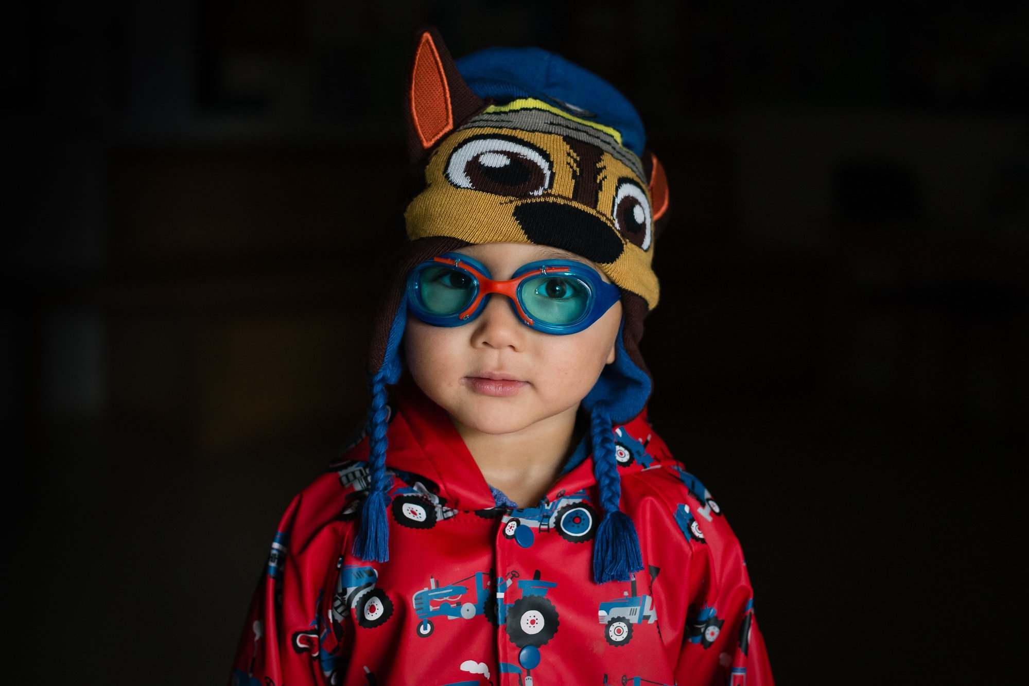 Young boy wearing a superhero costume with a red jacket featuring tractors, a blue swim cap, and aqua goggles, also wearing a brown and yellow animal-themed knit hat with large eyes and ears.
