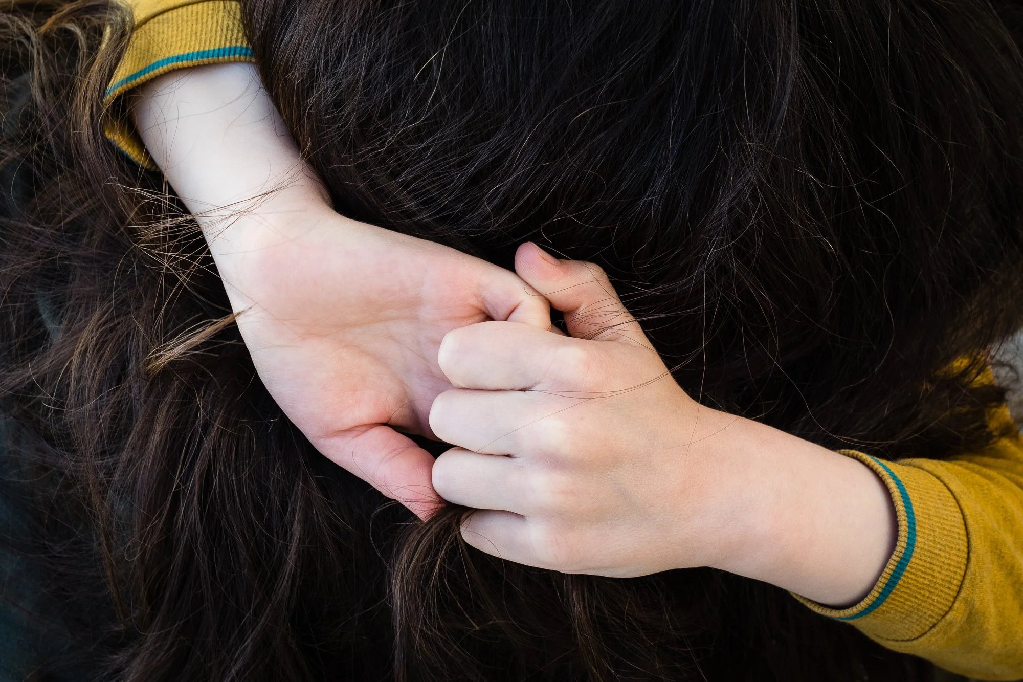 Person with dark hair, wearing a mustard yellow long sleeve shirt, holding a lock of hair.