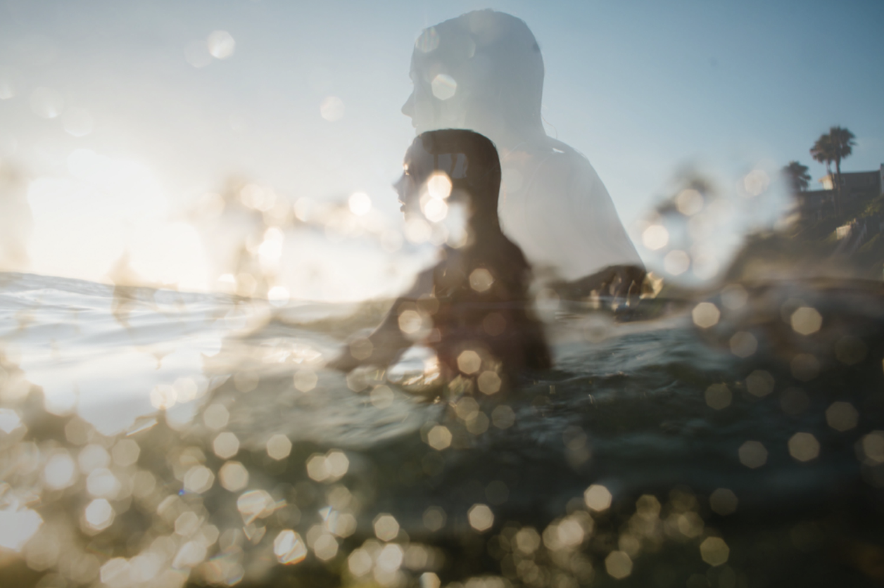 Two women swimming in the ocean during sunset, with one woman in the foreground and the other behind, with sunlight reflecting on the water and a house with palm trees in the background.