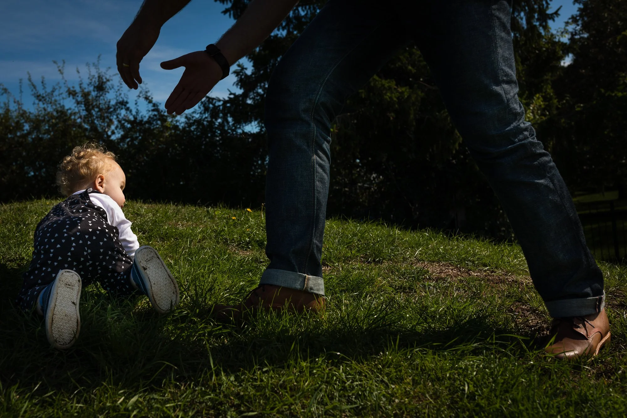 A person leaning forward with extended arms, seemingly playing with or helping a small child who is crawling on the grass outdoors during daytime.