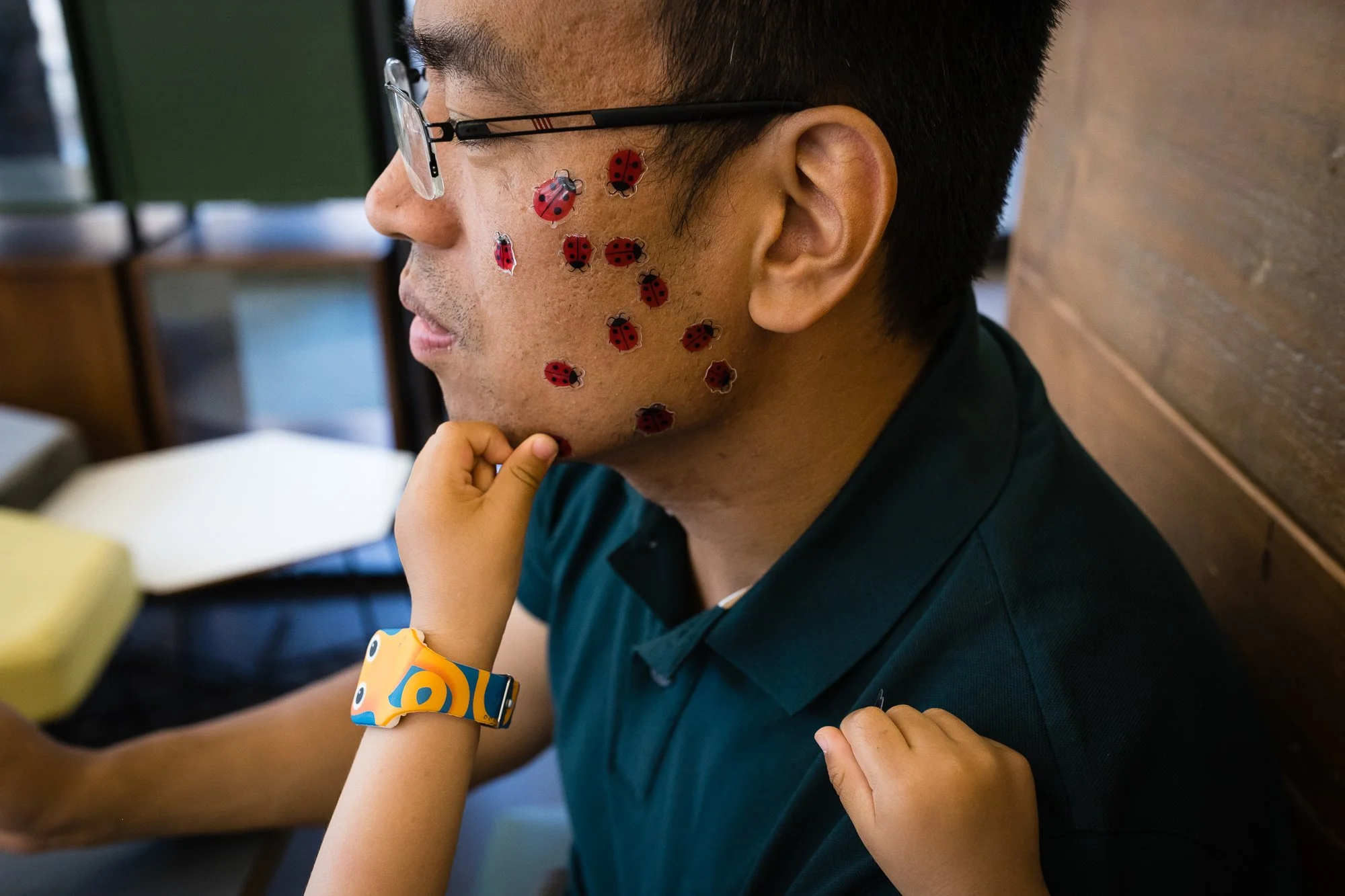A man wearing glasses with ladybug stickers on his face is sitting with a child touching his chin and arm, in an indoor setting.
