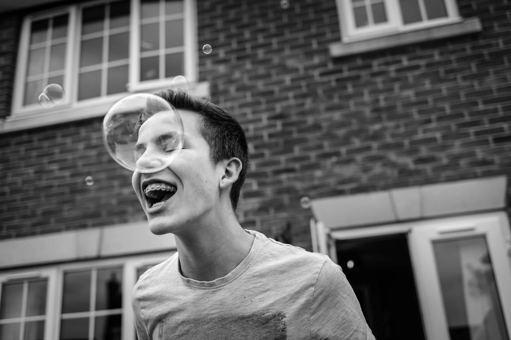 A young man with braces joyfully laughing while playing with soap bubbles outside a brick building.