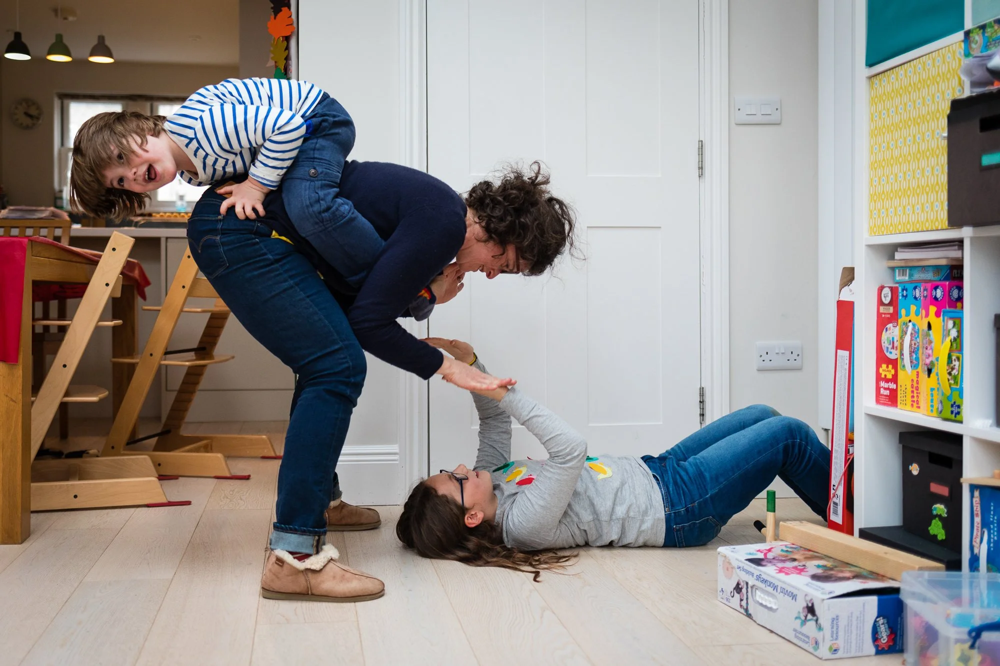 A woman lying on the floor in a playroom, lifting a young girl by the hands while a young boy on her back smiles and clasps his hands around the woman's waist.