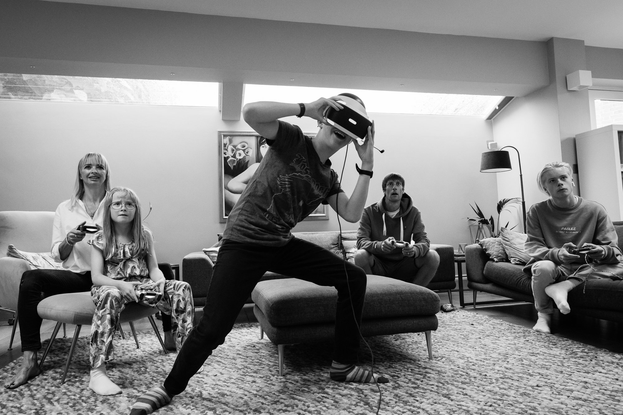Five people playing a virtual reality game in a living room, with some sitting on couches and others standing, holding controllers, and wearing VR headsets.