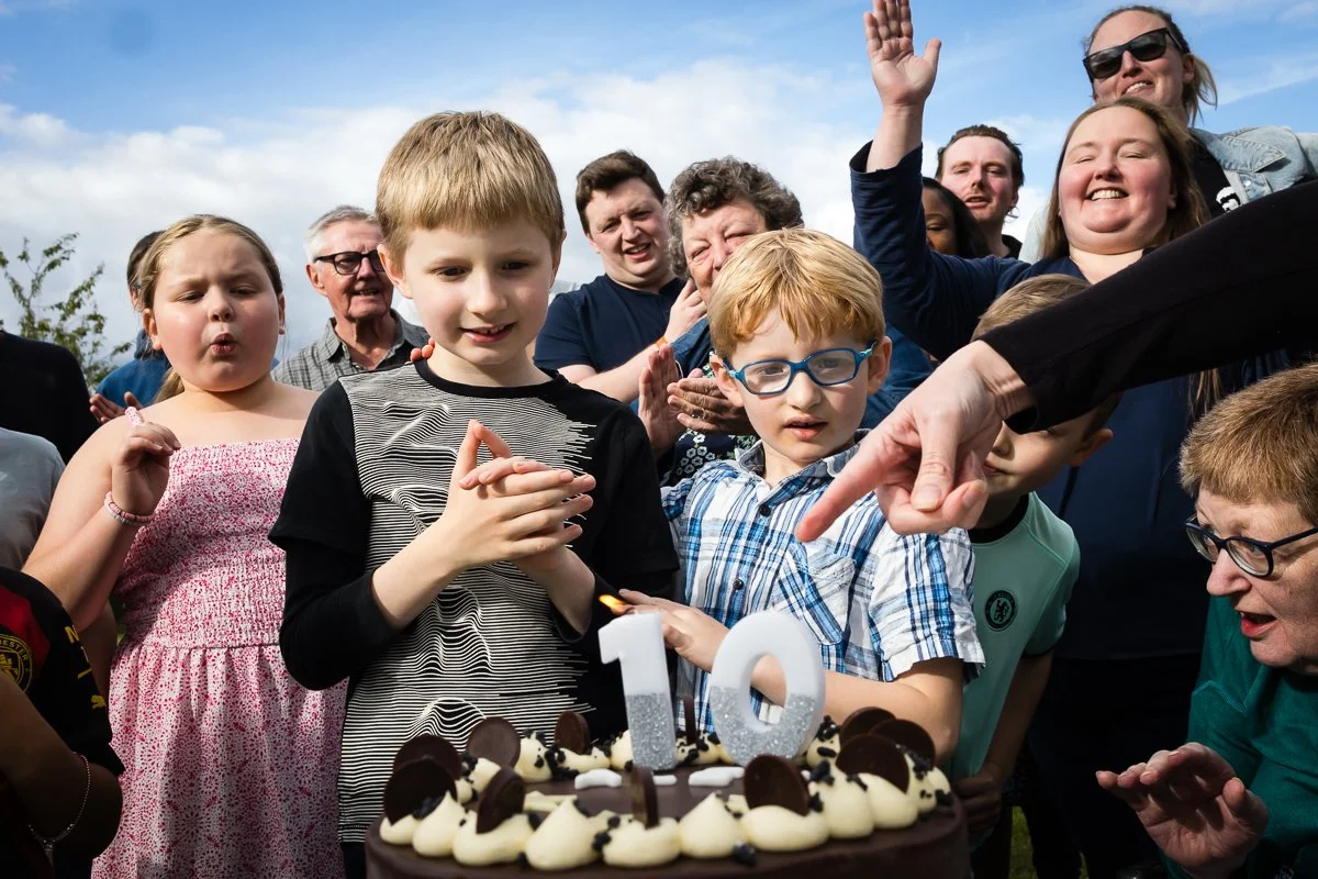 Group of children and adults celebrating a 10th birthday outdoors with a chocolate cake featuring a lit candle and the number 10 candle holder, many smiling and cheering.