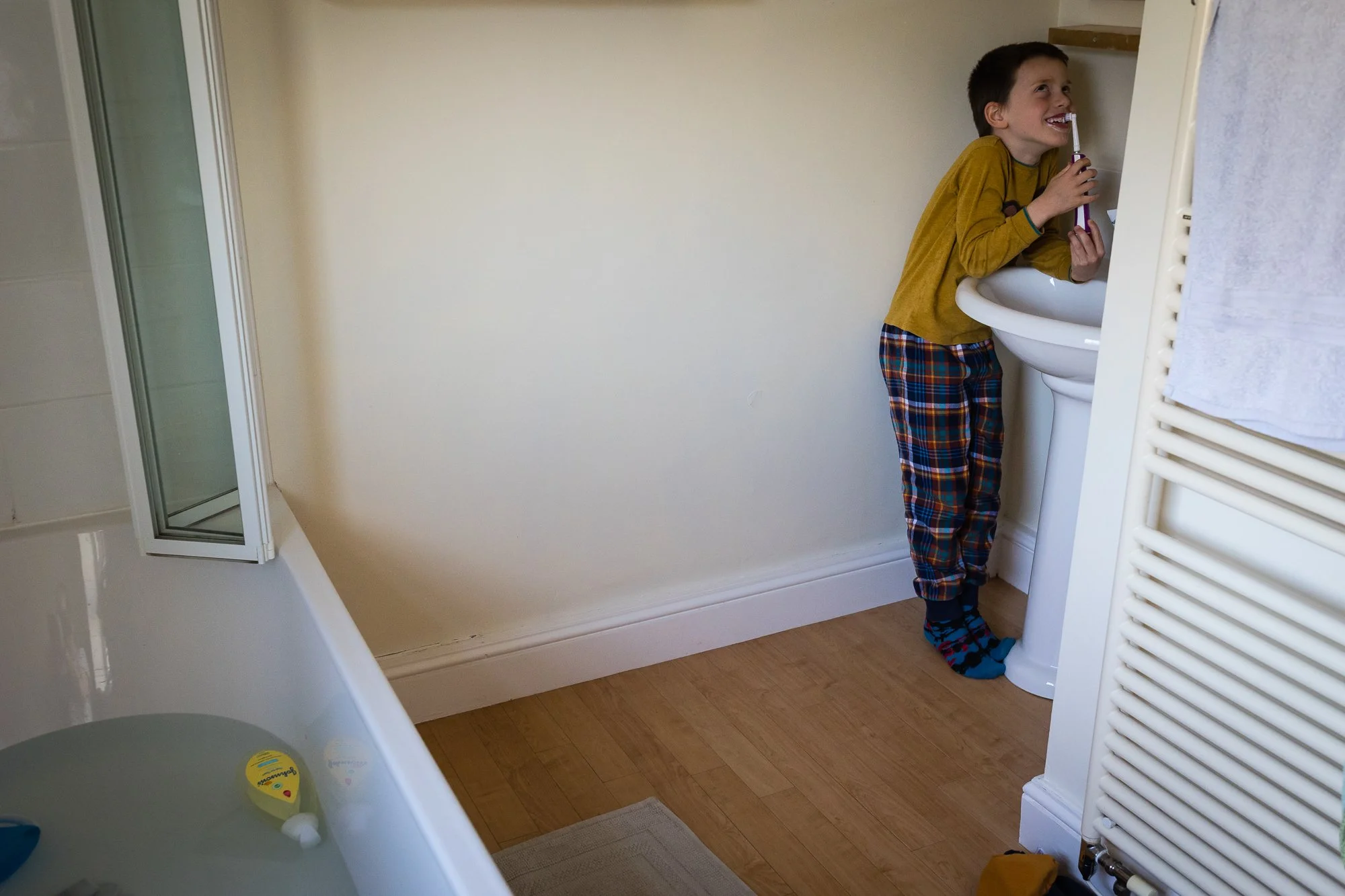 A young boy with dark hair, wearing a yellow long-sleeve shirt and plaid pajama pants, brushing his teeth at a bathroom sink, smiling and looking up.