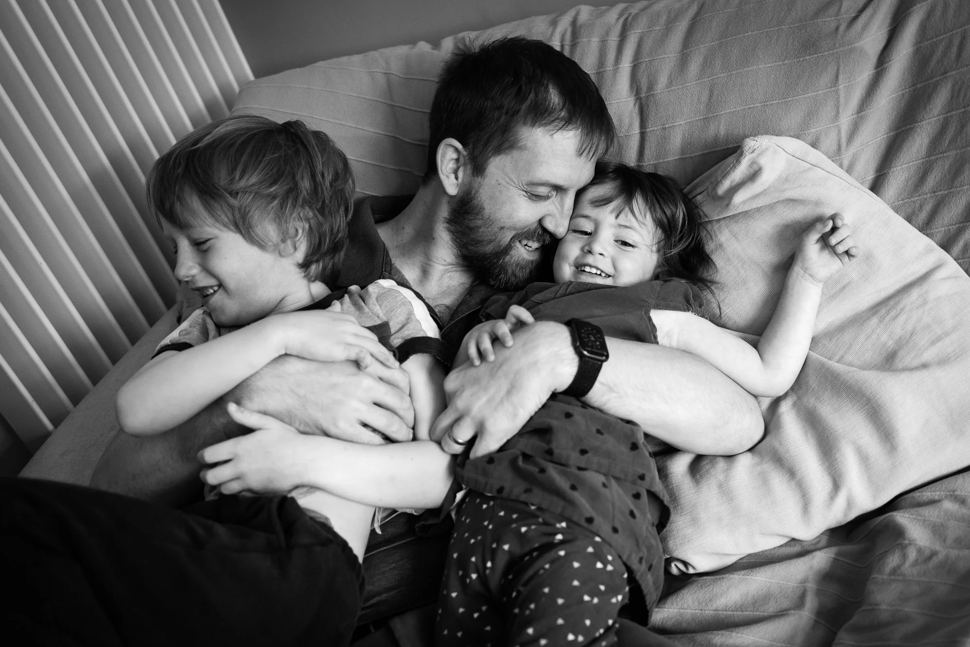 A man and two children, a boy and a girl, sharing a joyful cuddle on a bed, smiling and hugging each other in a moment of happiness.