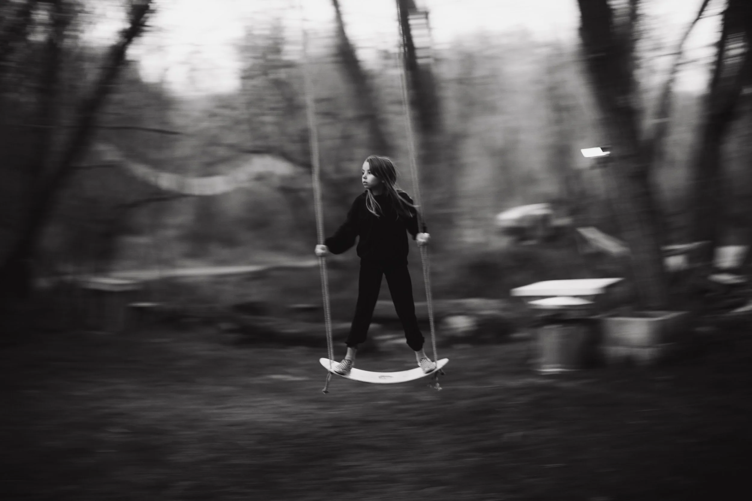 Black and white photo of a young girl swinging on a swing set outdoors with trees in the background.