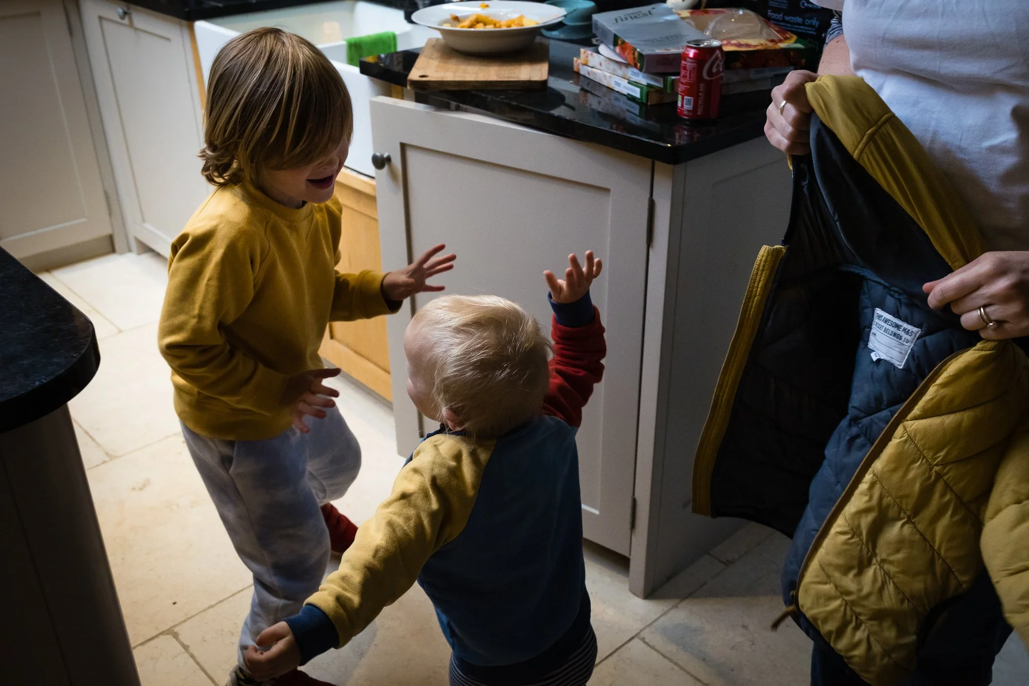 Two young children in a kitchen, one with brown hair and wearing a yellow sweatshirt, appearing upset, and the other with blond hair, wearing a red and blue jacket, reaching up towards an adult holding a yellow jacket. The kitchen countertop in the b