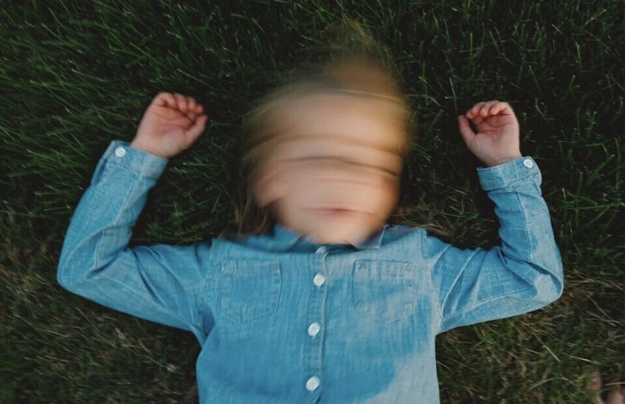A young girl lying on her back on grass with her arms raised, wearing a denim jacket, her face blurred due to motion.
