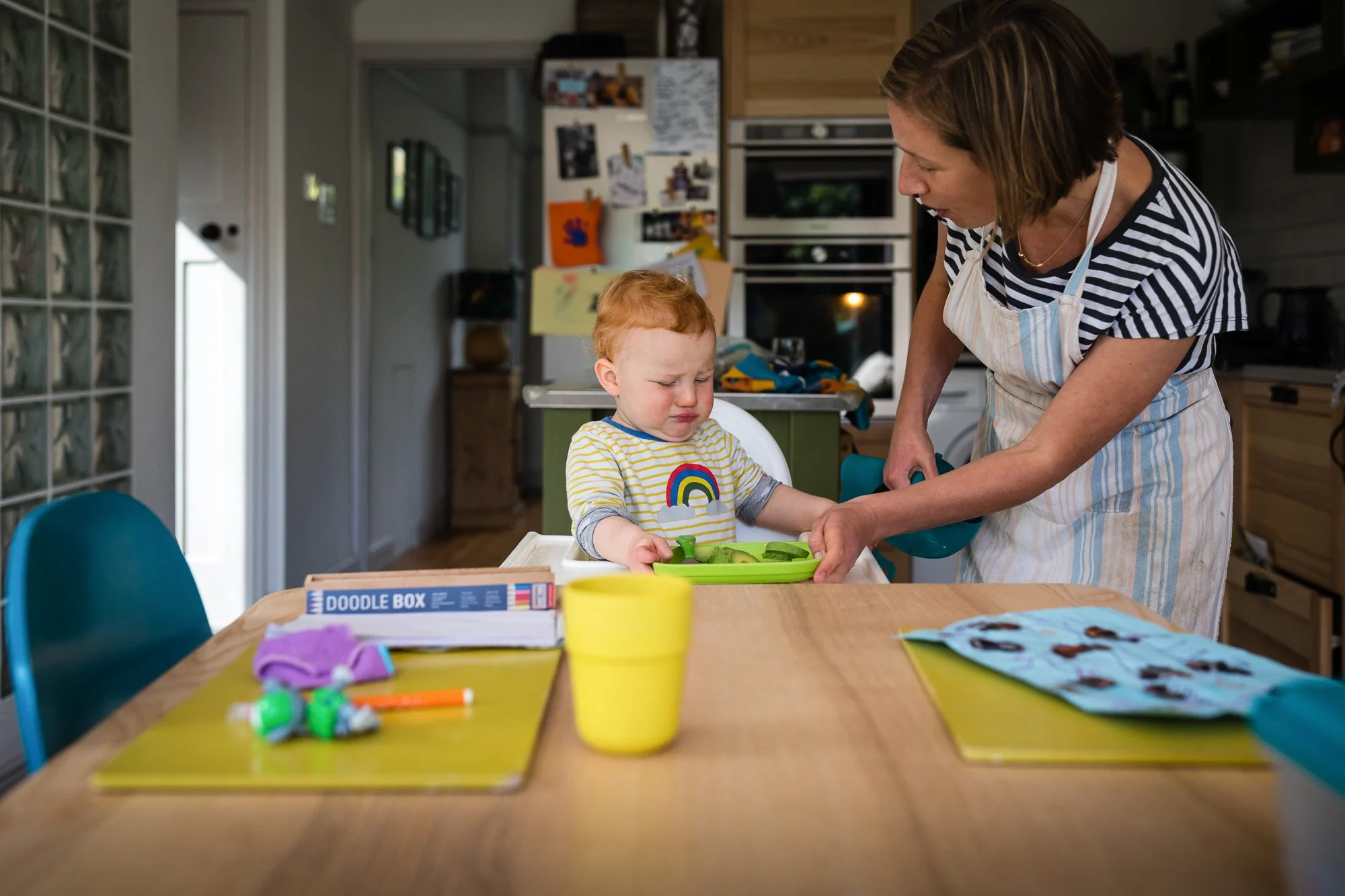 A young child with red hair and a rainbow rainbow shirt sitting at a dining table, looking upset, as an adult woman, possibly his mother, feeds him vegetables from a green tray. The setting is a kitchen with various items on the table and in the back