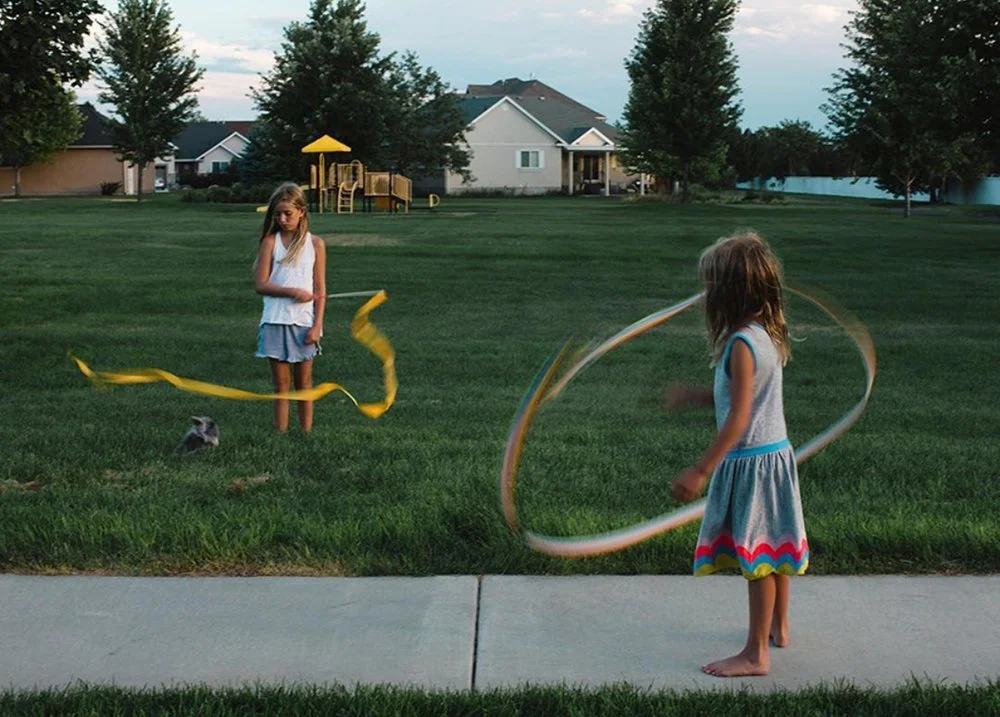 Two young girls playing with hula hoops on a sidewalk in a grassy park area, with houses and trees in the background during dusk.