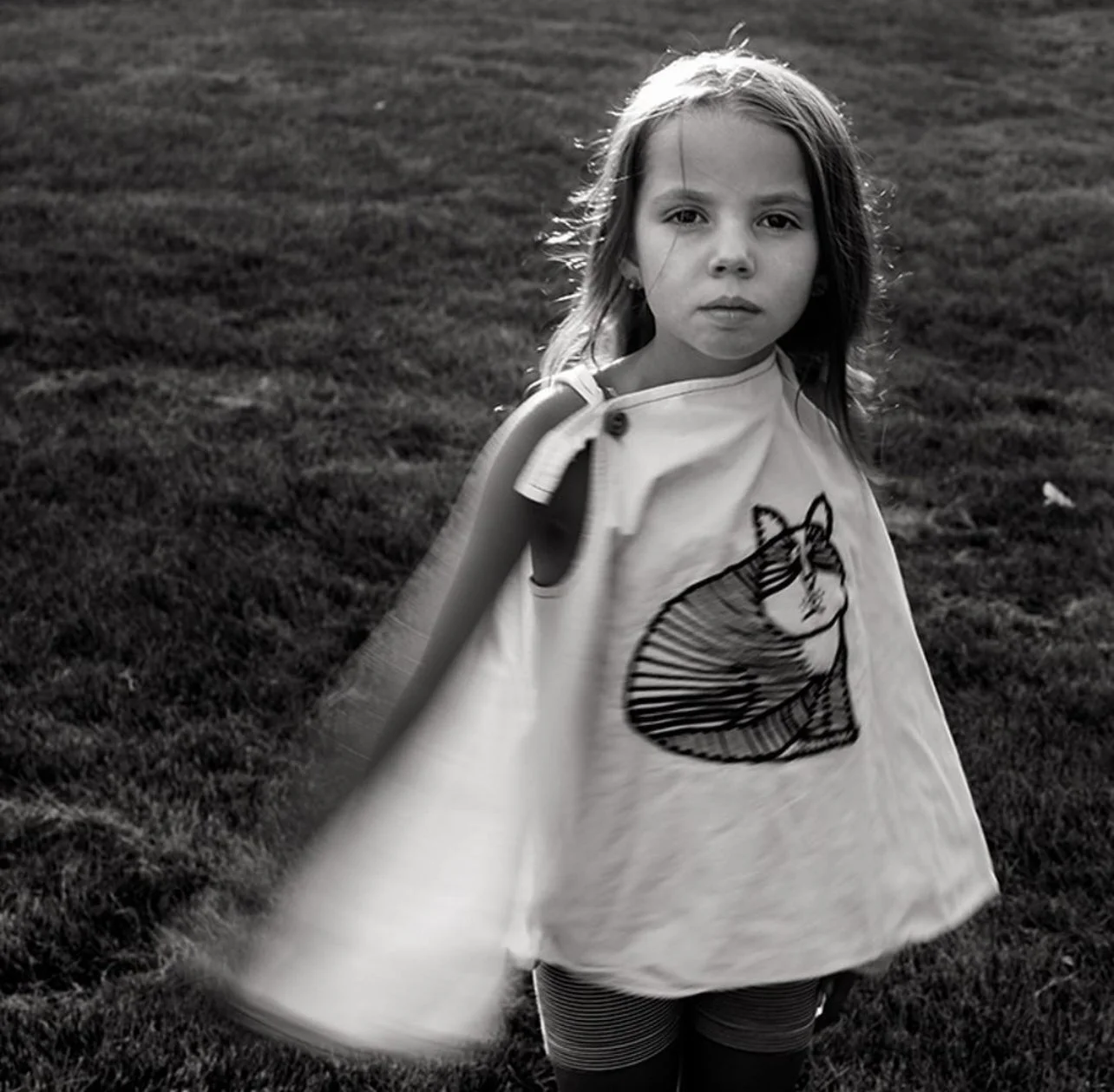 Black and white photo of a young girl with long hair standing on grass, wearing a cape with a cat illustration and striped leggings, looking directly at the camera.