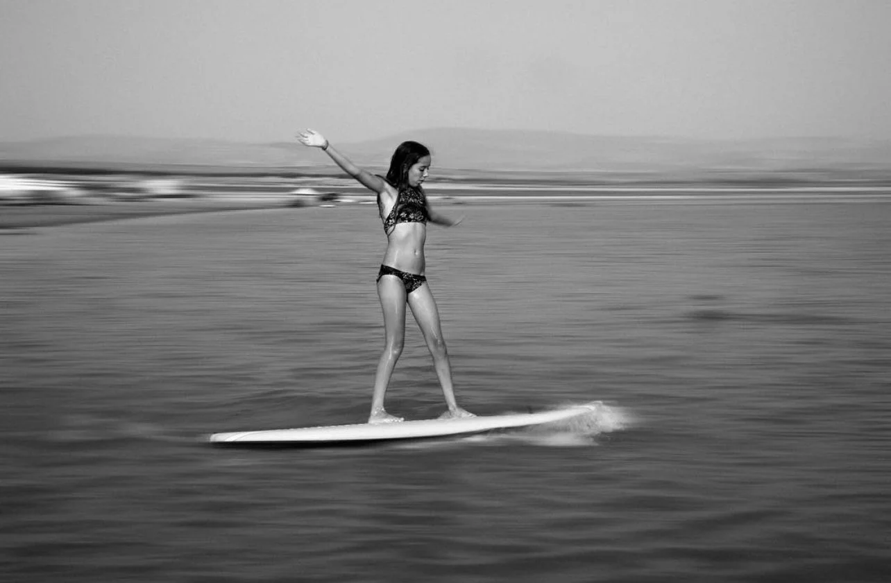 A woman in a bikini standing on a surfboard in the water, with her arms outstretched for balance.