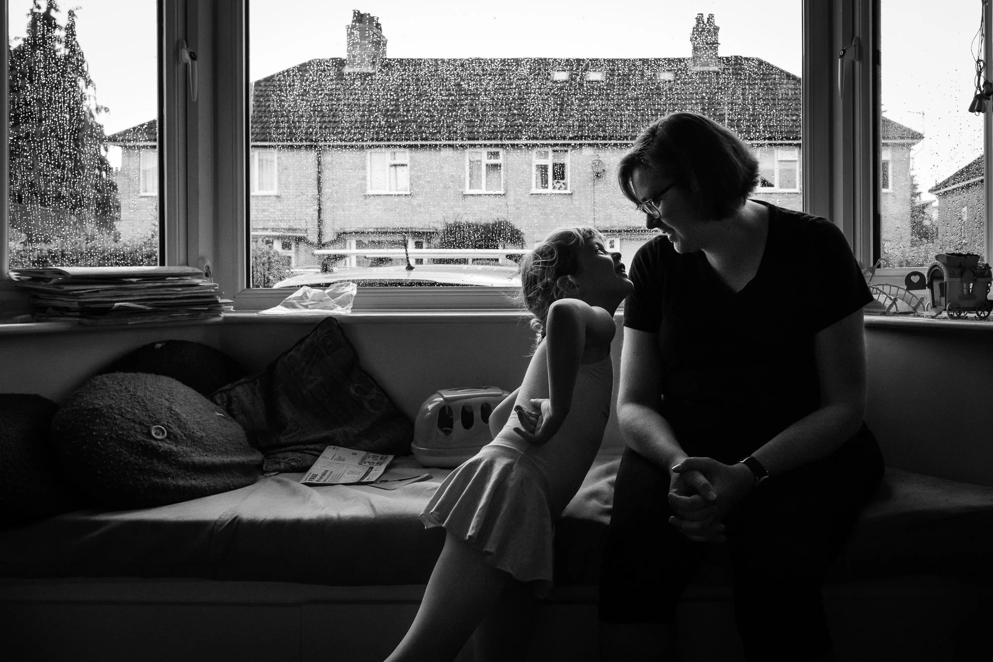 A woman and a young girl sitting on a window seat indoors, with rain on the window behind them, sharing a joyful moment and looking at each other.