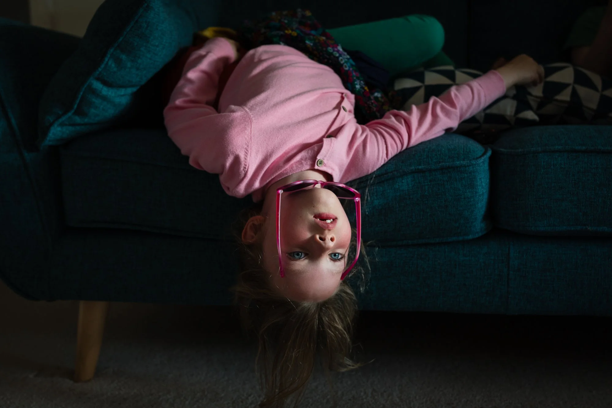 A young girl with blonde hair and blue eyes is lying upside down on a dark teal couch, wearing pink sunglasses, a pink jacket, and looking at the camera. The couch has patterned pillows and is near a dark background.