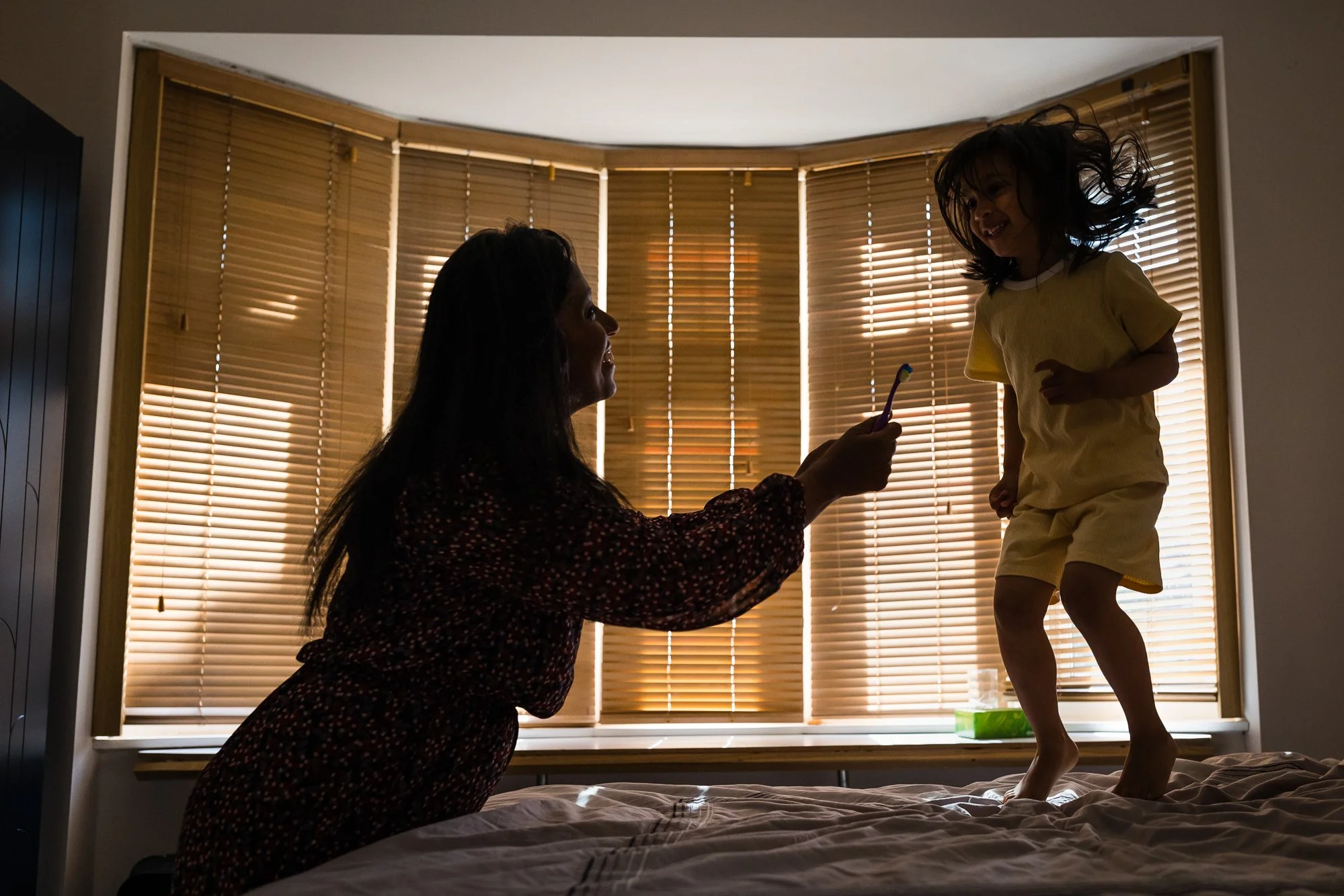 A woman kneeling on a bed holding a toothbrush towards a young girl jumping on the bed, with wooden blinds in the background.