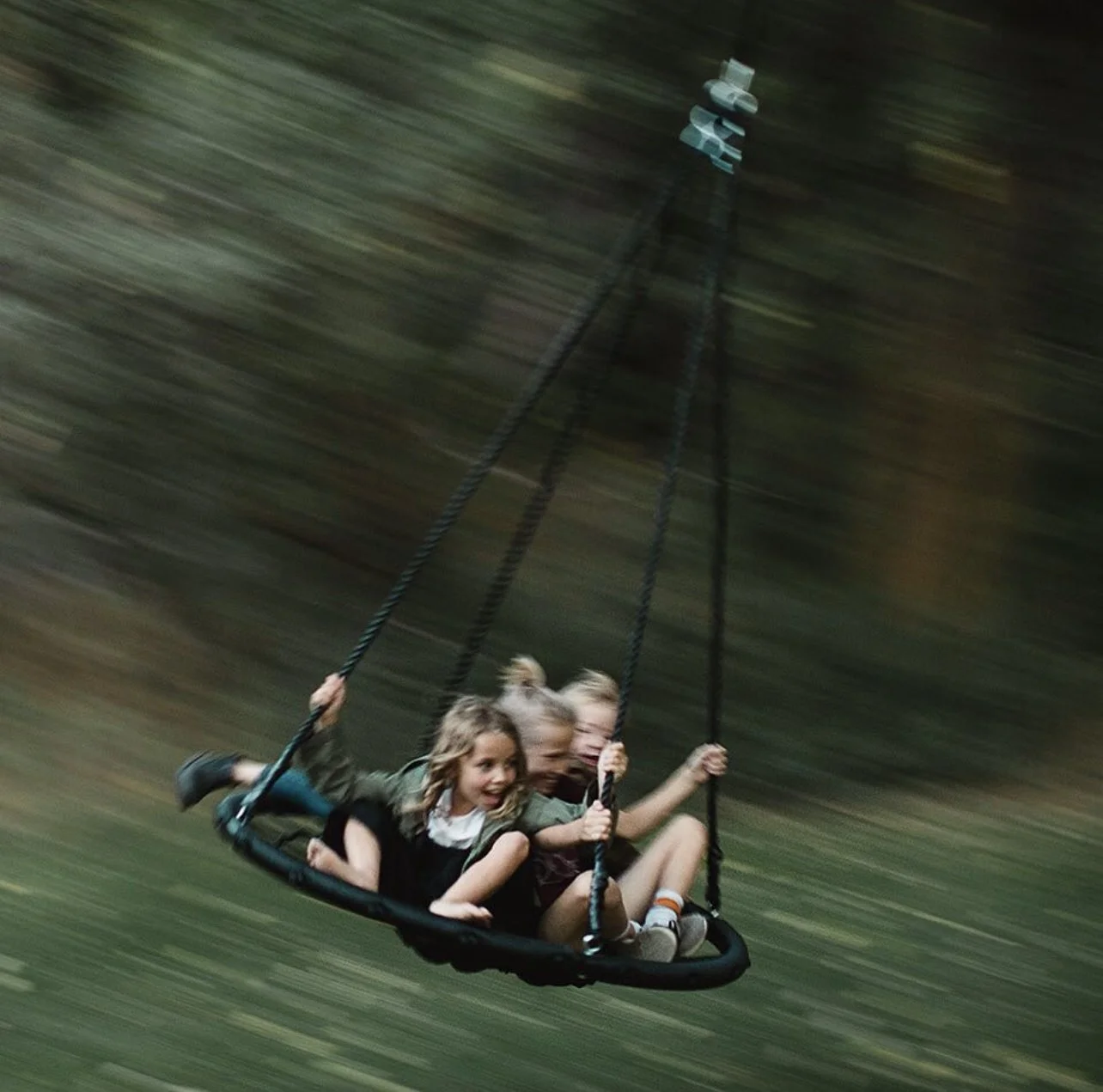 Three children on a swing hanging from a metal frame, swinging quickly through a blurred outdoor background.