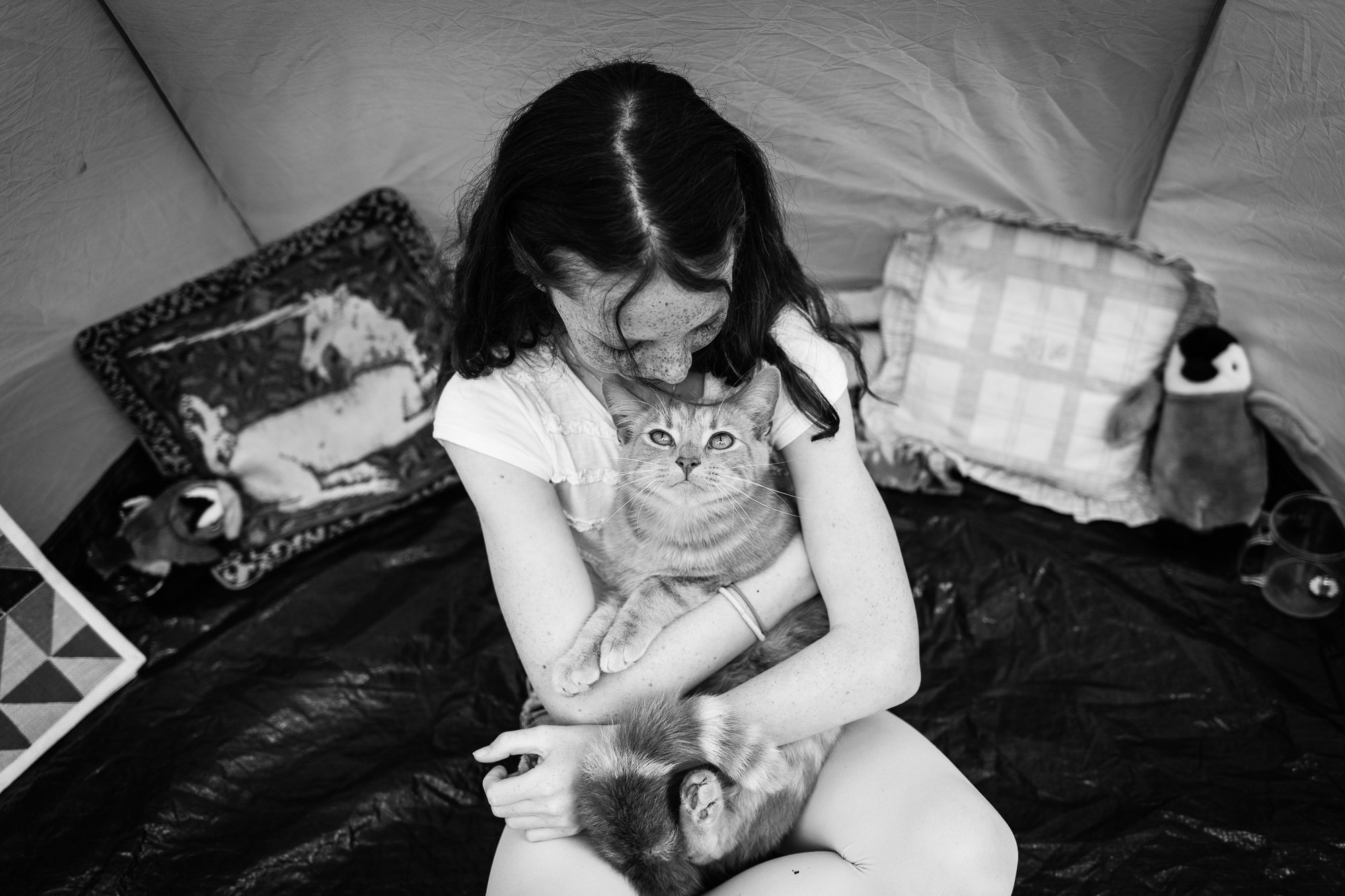 A girl with curly hair and freckles sitting inside a tent, hugging two cats, one orange and one with darker fur, looking directly at the camera.