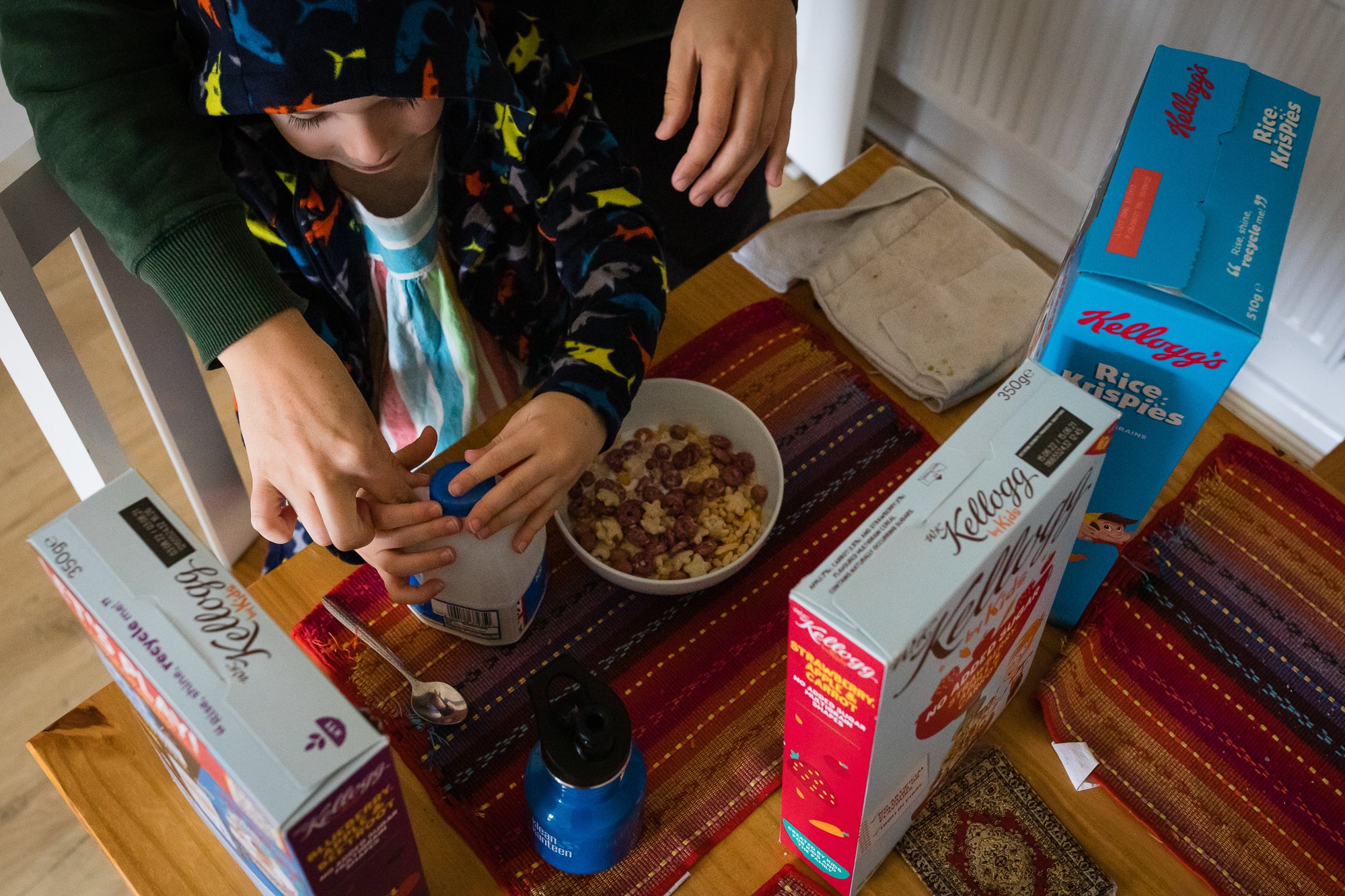 Child opening a cereal box at a table with cereal bowls and additional cereal boxes on the table.