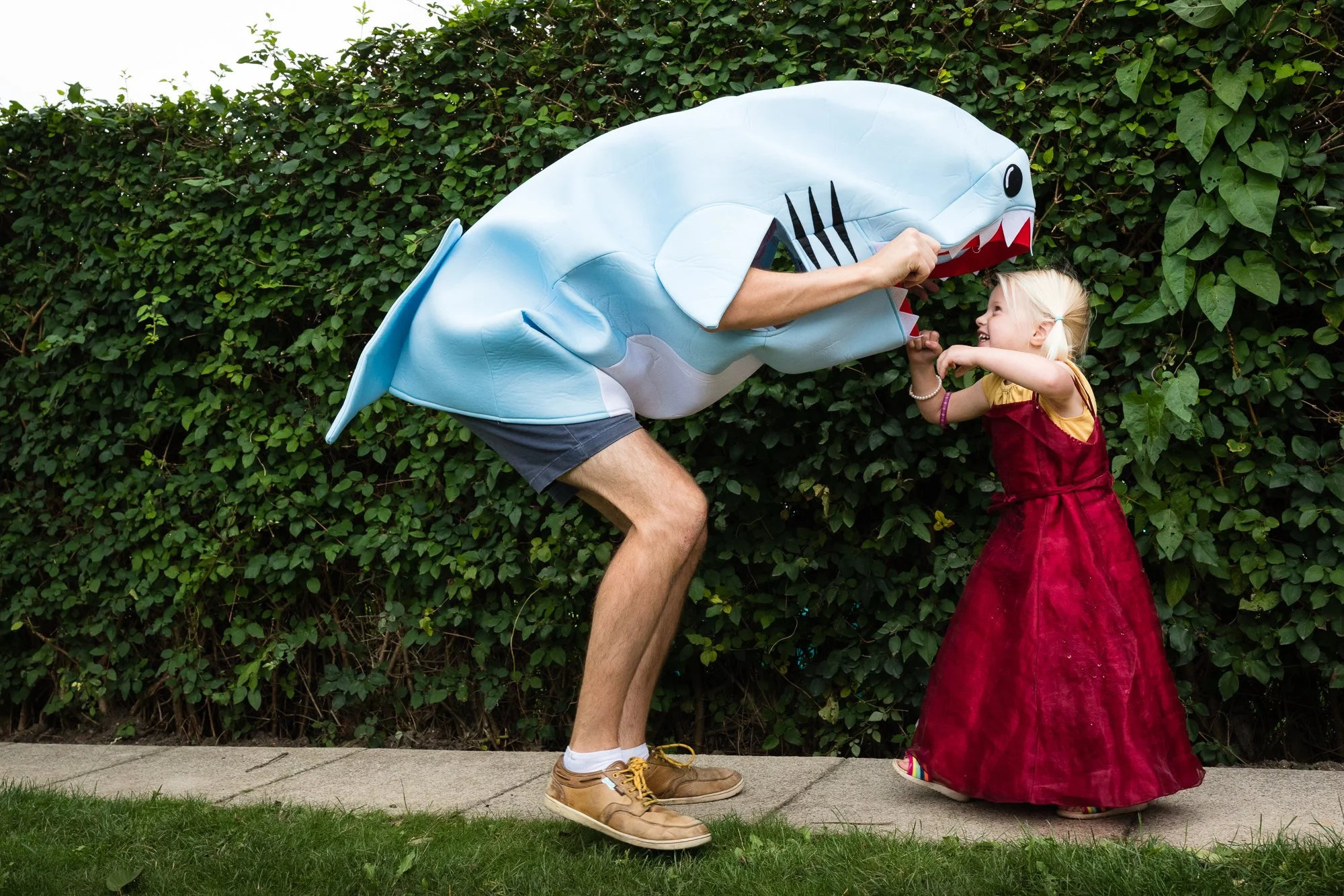 Person in a shark costume playing with a young girl in a red dress outside on a sidewalk with green bushes in the background.