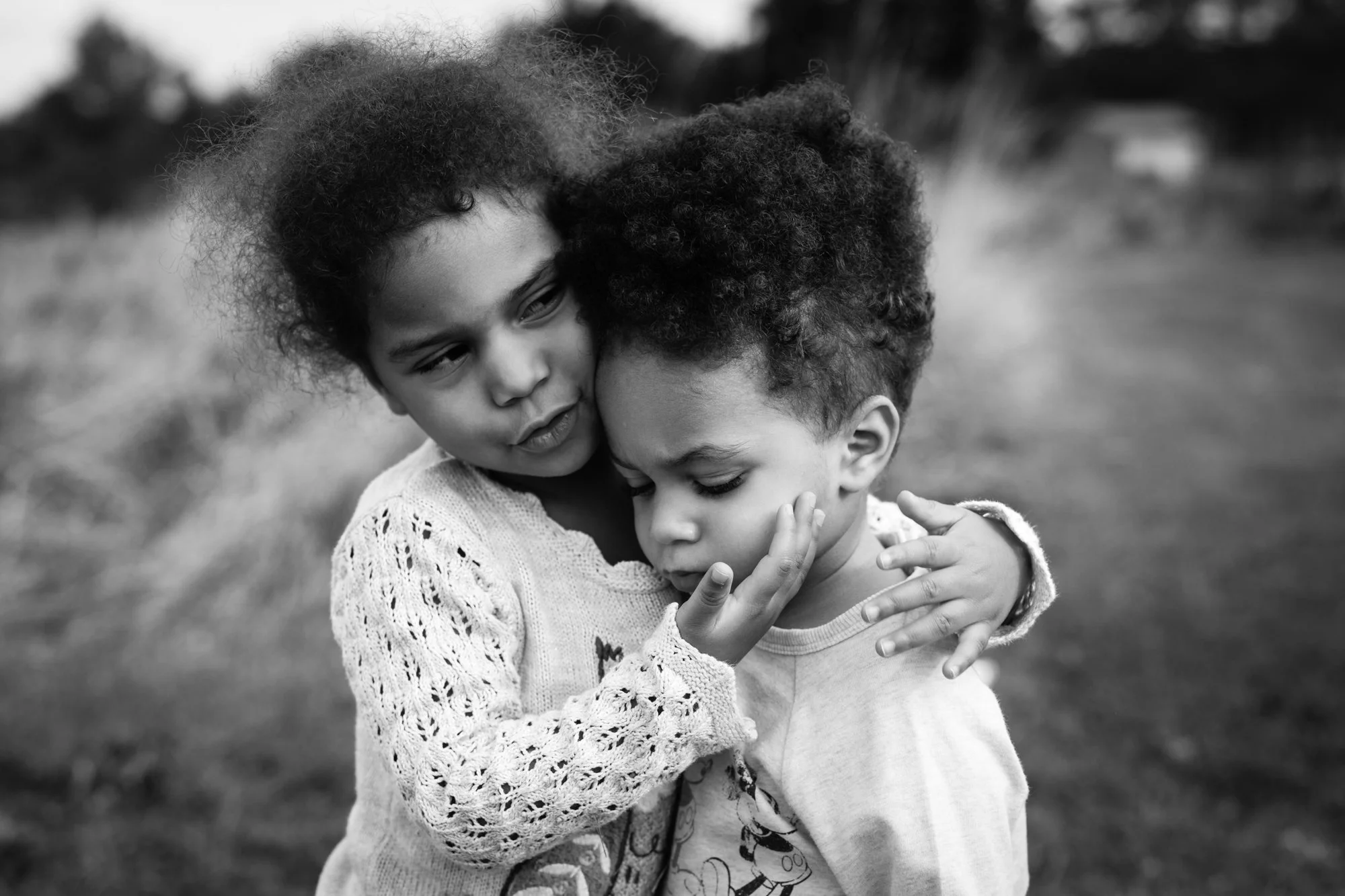 Two young children hugging outdoors, one with curly hair and the other with frizzy hair, in black and white.