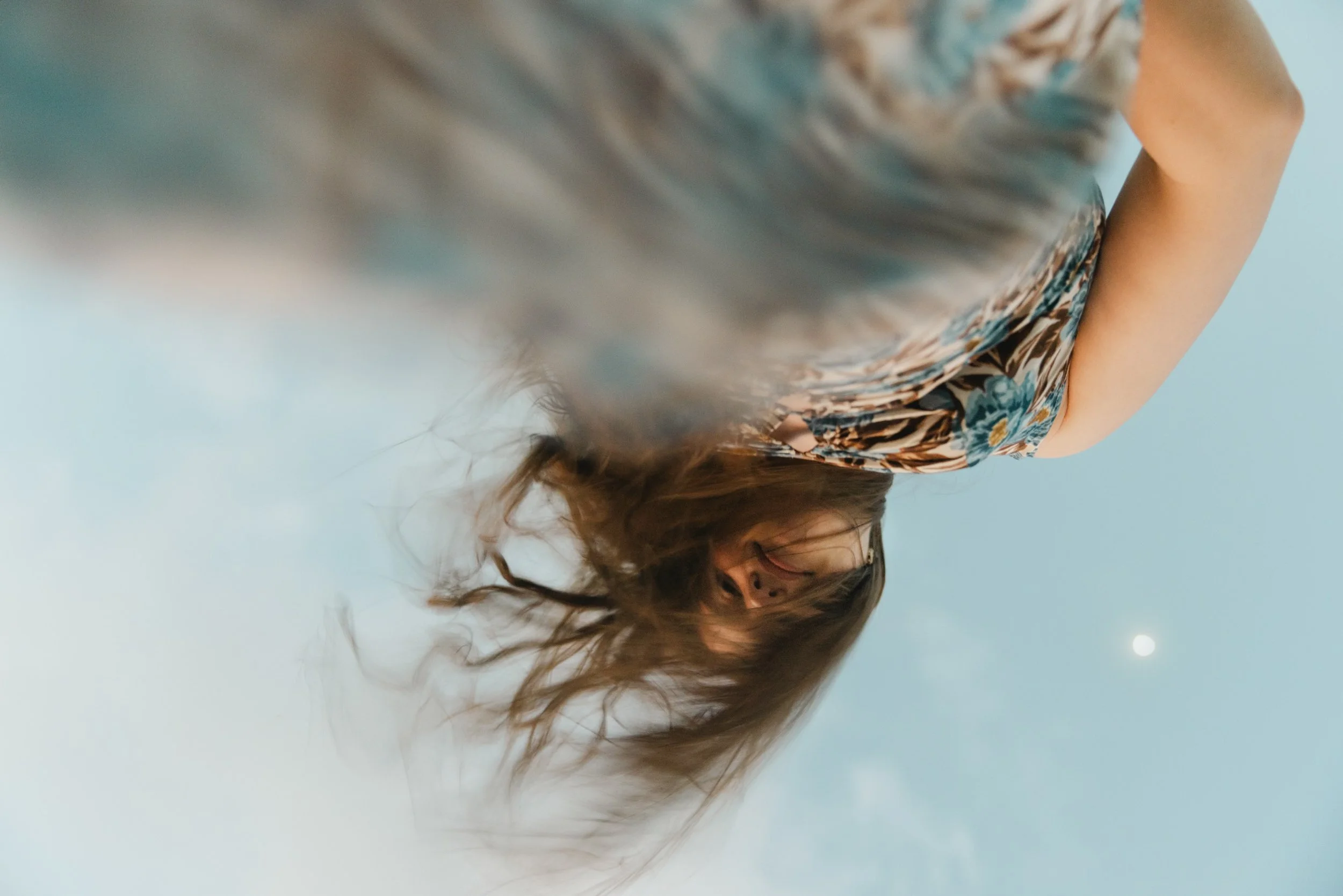 A woman with long hair and glasses looking at the camera, wearing a floral dress, against a cloudy sky with the moon visible in the background.