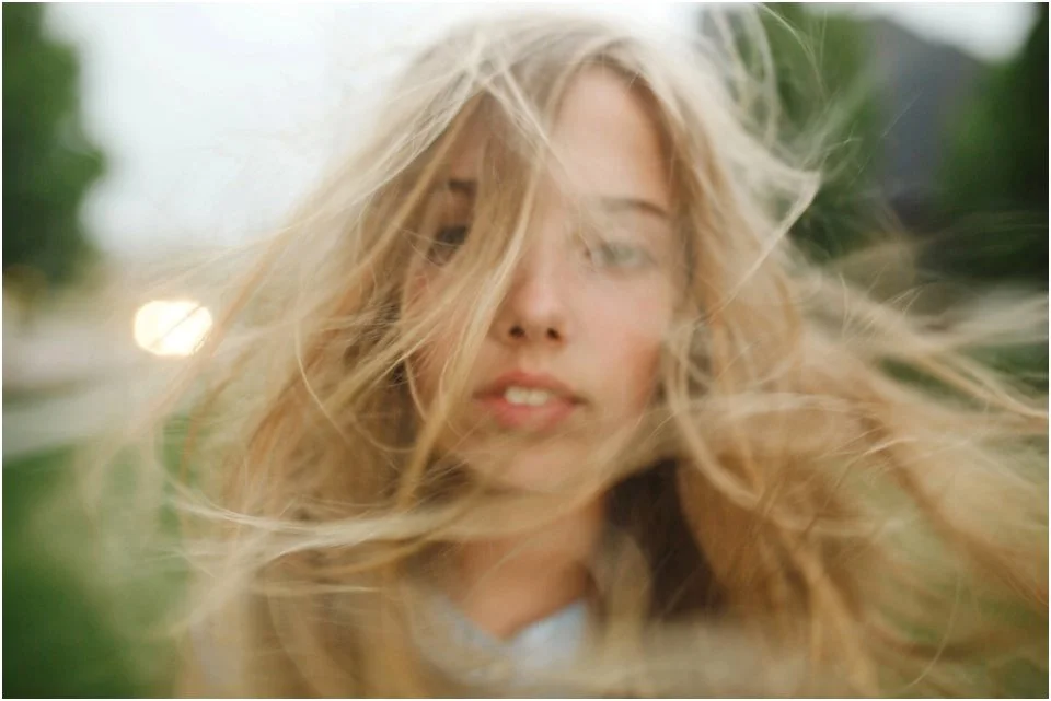 A young woman with long blonde hair outdoors, slightly blurred, with a soft natural background.