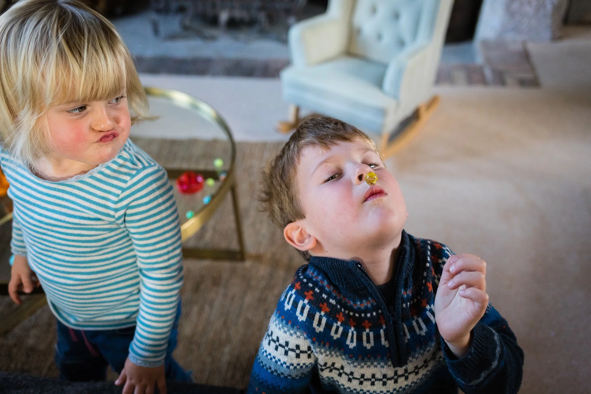 Two children, a girl with blonde hair in a striped shirt and a boy in a patterned sweater, are playing with colorful capsules or candies indoors near a table with a glass top and toys.