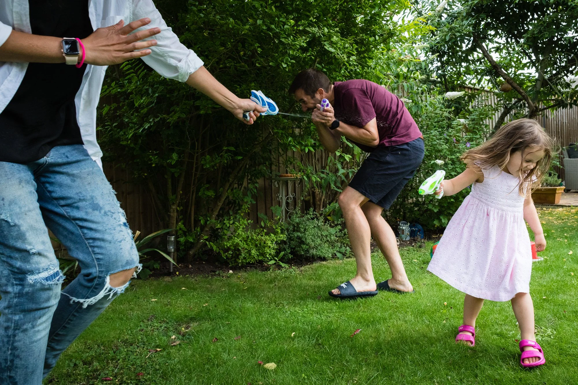 A family in a garden engaged in water gun fight, with a woman crouching and a girl holding a water gun, surrounded by greenery.